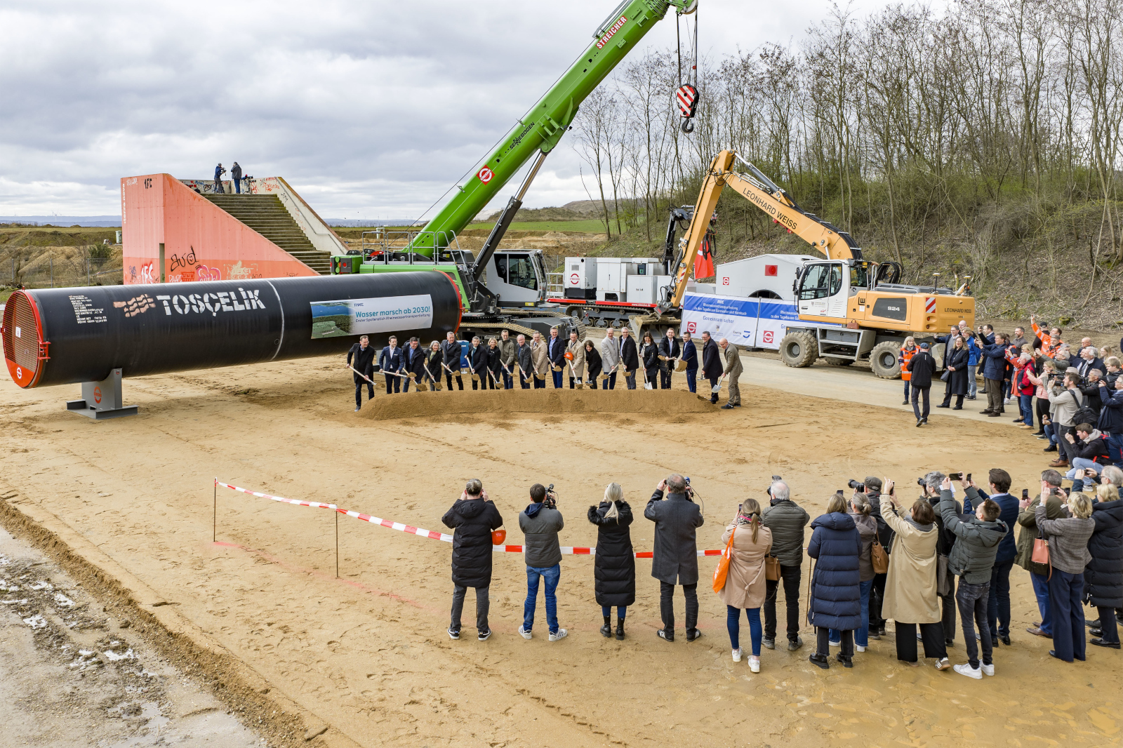 Politiker und Vertreter führen eine Grundsteinlegung vor einem groÃen schwarzen TOSCELIK-Rohr durch, umgeben von Kranen, Baggern und Fotografen.