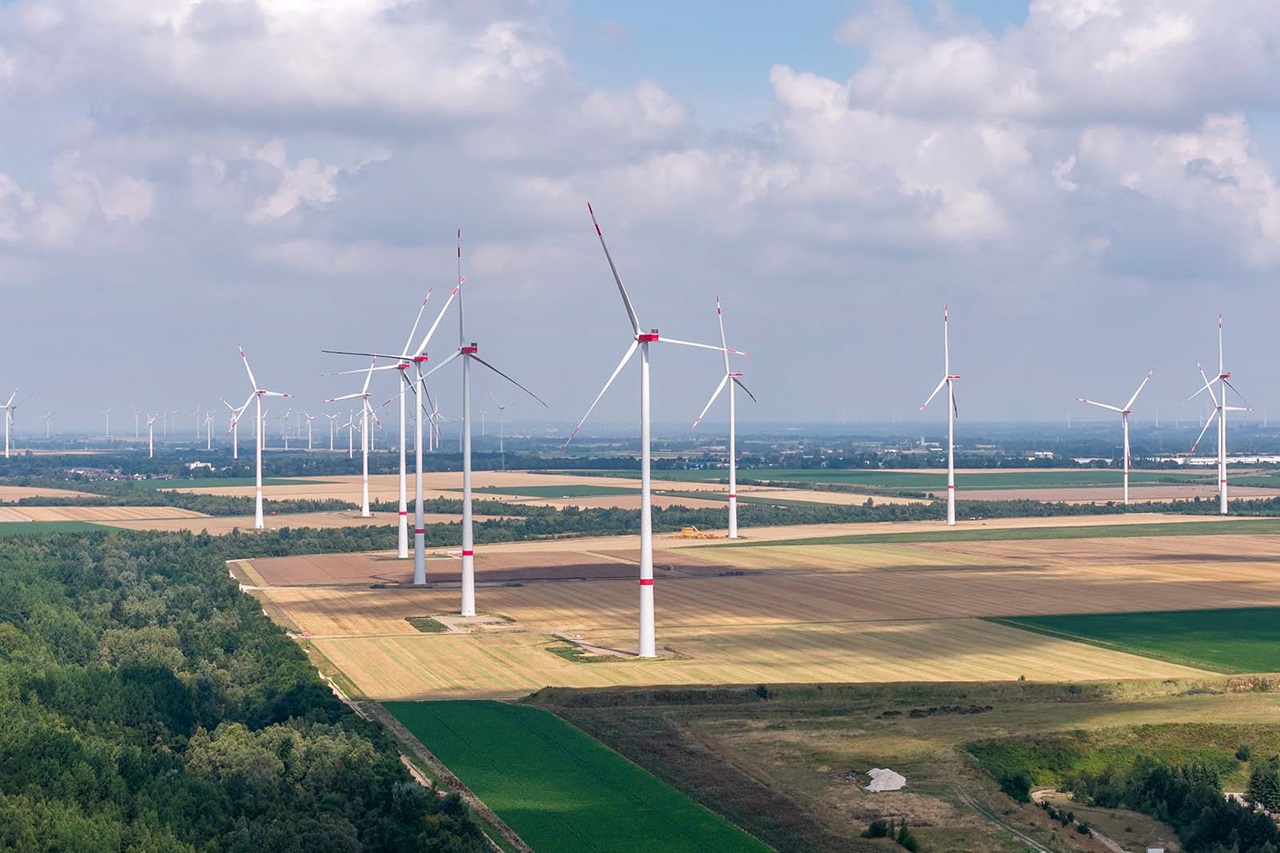 Mehrere moderne Windräder stehen auf weitläufigen Feldern neben einem Wald; die Rotorblätter sind rot markiert und werfen Schatten, der Himmel ist wolkig.