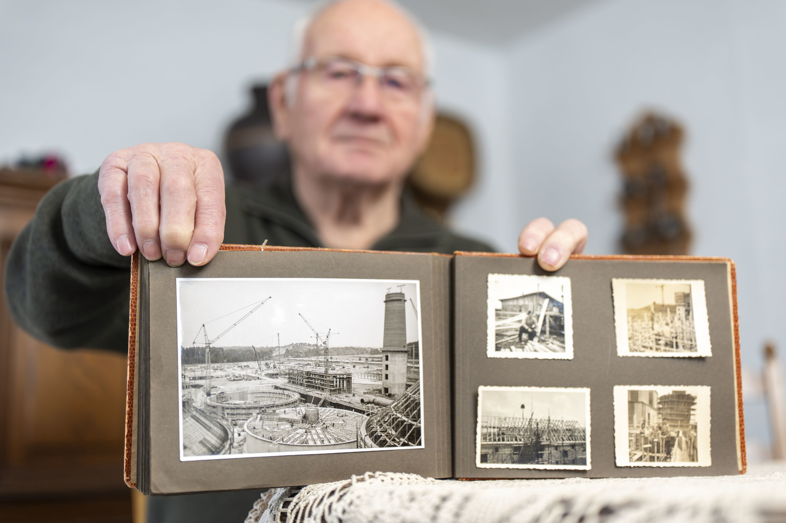 Eine Person hält ein altes Fotoalbum mit Schwarzweissaufnahmen einer Baustelle, mit Kränen, runden Becken und vier kleinen Fotos rechts.