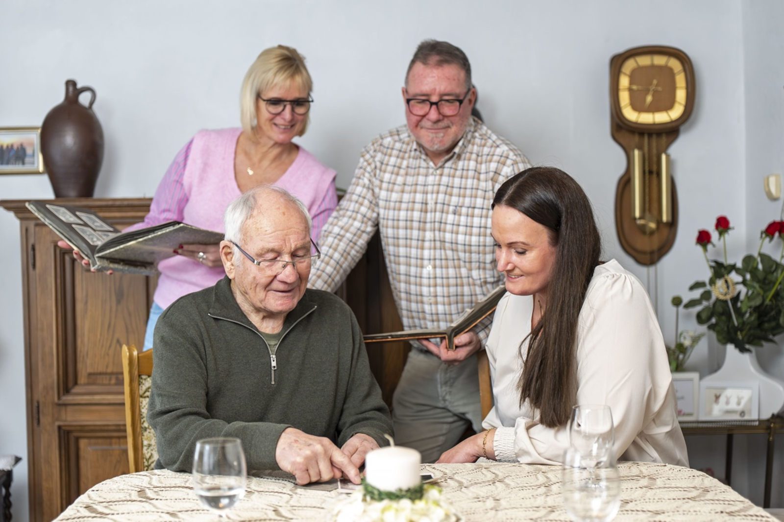 Vier Familienmitglieder mit unkenntlich gemachten Gesichtern sitzen und stehen um einen Tisch mit Häkeldecke, Kerze, Trinkgläsern, Rosen und Fotoalben.