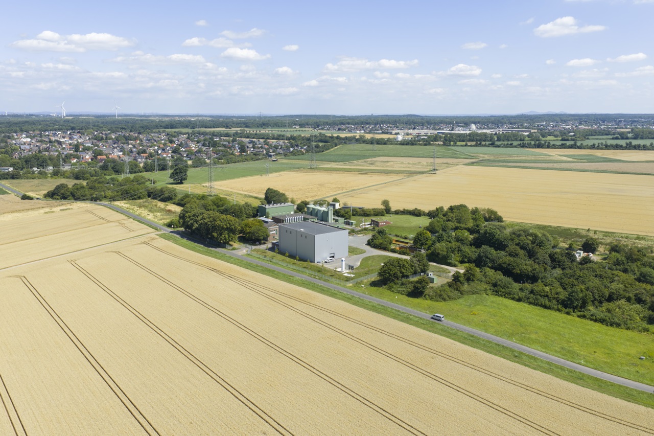 Eine graue Lagerhalle mit glatter Metallfassade steht im Bildvordergrund zwischen goldenen Getreidefeldern und grünen Baumgruppen. Eine schmale Landstraße mit einem Auto verläuft davor; im Hintergrund liegen ein Dorf und Felder unter hellem blauem Himmel.
