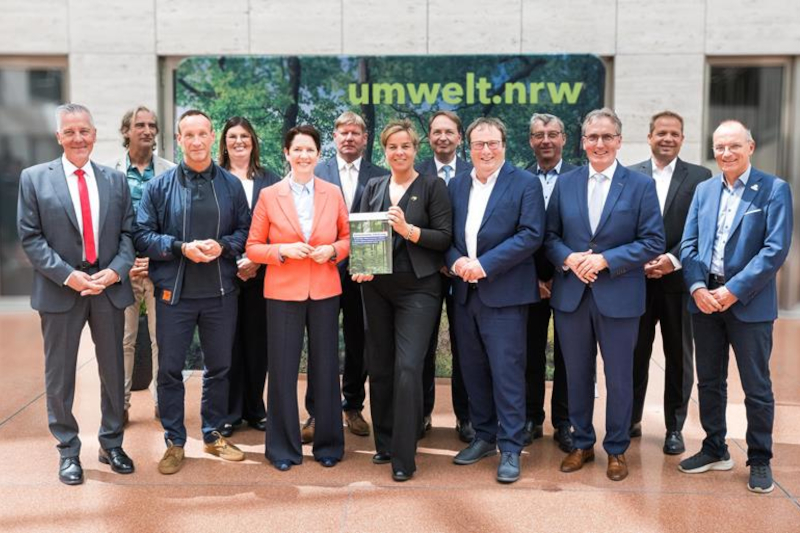 Eine Gruppe von Personen in dunklen Anzügen und einer orangefarbenen Jacke steht vor einem grünen Banner mit dem Text „umwelt.nrw“ in einer hellen Marmorlobby.