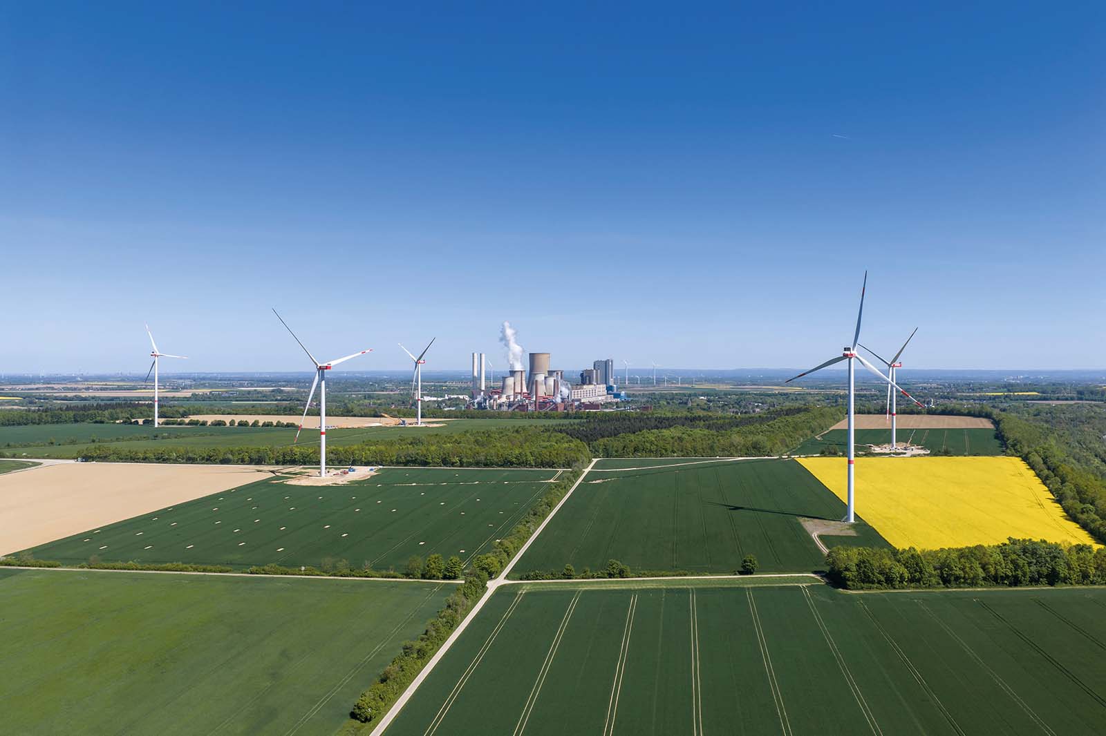 Mehrere weiße Windturbinen mit metallenen Türmen ragen über rechteckige, grüne und gelbe Ackerflächen, im Hintergrund ein Industriekomplex mit rauchenden Kühltürmen unter klarem blauem Himmel.