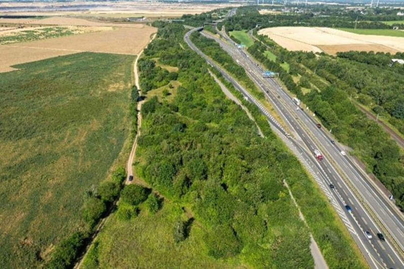 Luftaufnahme einer breiten Autobahn mit mehreren Fahrspuren und Fahrzeugen rechts, daneben ein breiter grüner Baumstreifen, links goldene und grüne Felder unter hellem Himmel.