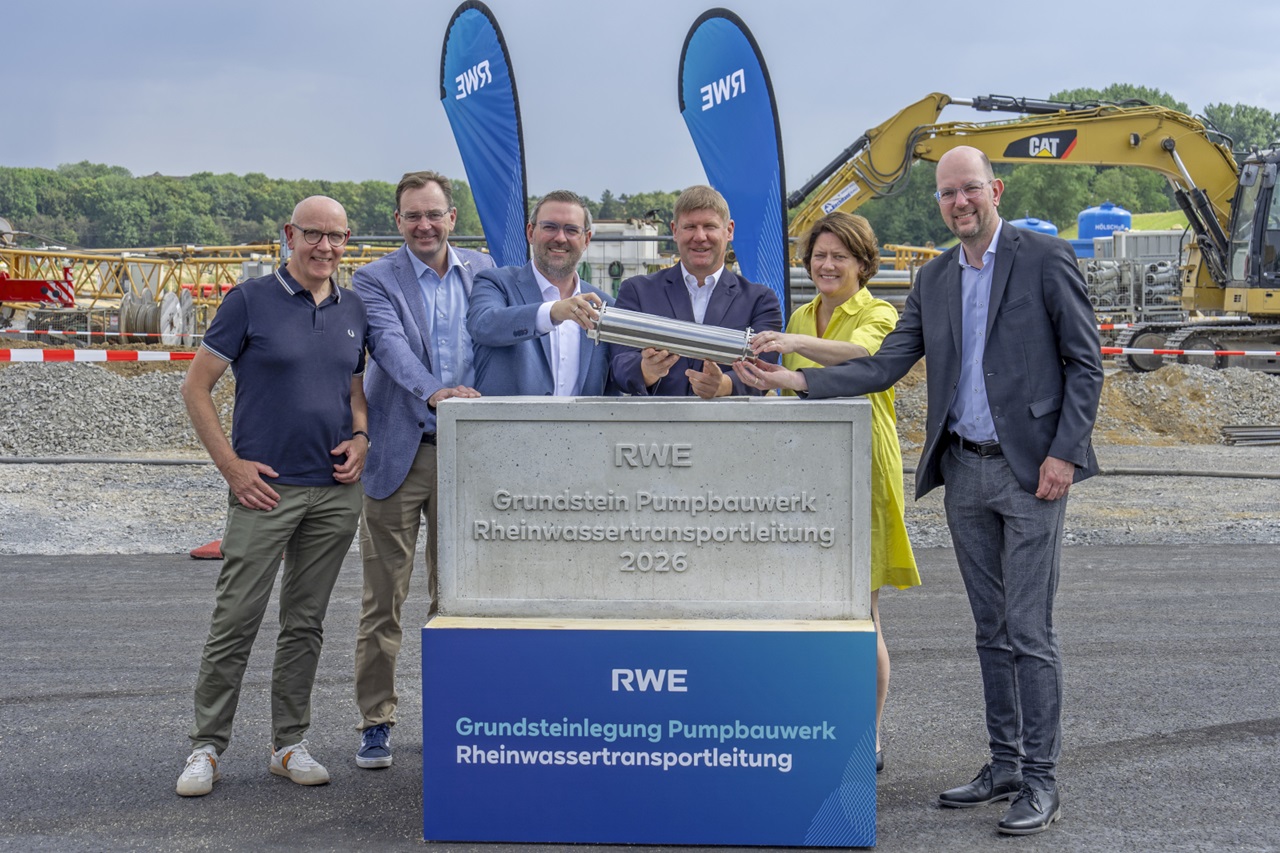Sieben lächelnde Personen halten gemeinsam eine silberne Metallkapsel über einen grauen Grundstein mit der Aufschrift „RWE“ und „Grundsteinlegung Pumpbauwerk Rheinwassertransportleitung“ vor einer Baustelle.