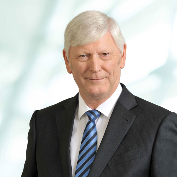 A man is wearing a black suit with a striped tie and has grey hair, set against a bright, blurred background, portrait of Dr. Rolf Martin Schmitz.