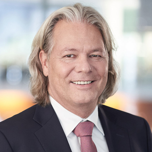 A man with long, wavy hair wearing a dark suit and a light blue shirt with a pink tie, set in a blurred background, portrait of Andree Stracke.