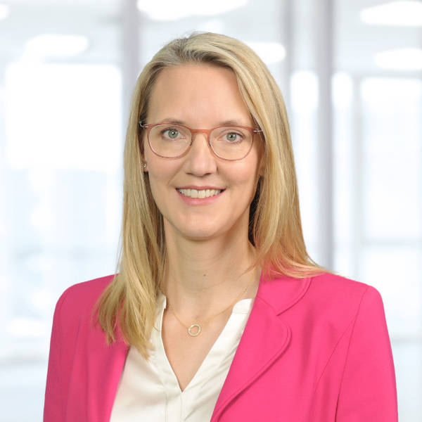 Eine Person mit blonden Haaren trägt ein pinkes Blazer und eine weiße Bluse. Der Hintergrund ist hell und unscharf, Portrait von Katja Wünschel.