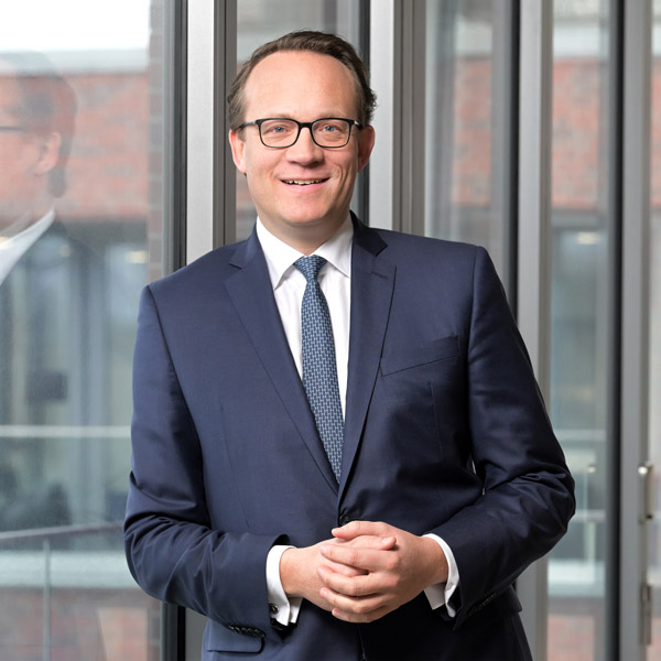 A person in a navy suit stands in front of large glass windows, hands clasped, with a blurred face, portrait of Dr Markus Krebber.