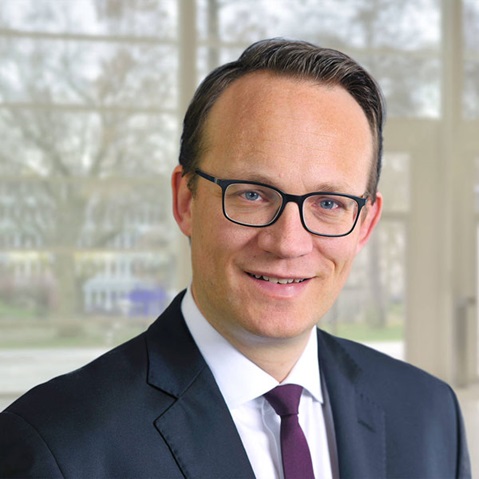 Ein Mann trägt einen Anzug mit einer Krawatte und steht in einem modernen, hellen Raum mit Fenstern im Hintergrund, Portrait von Dr. Markus Krebber.