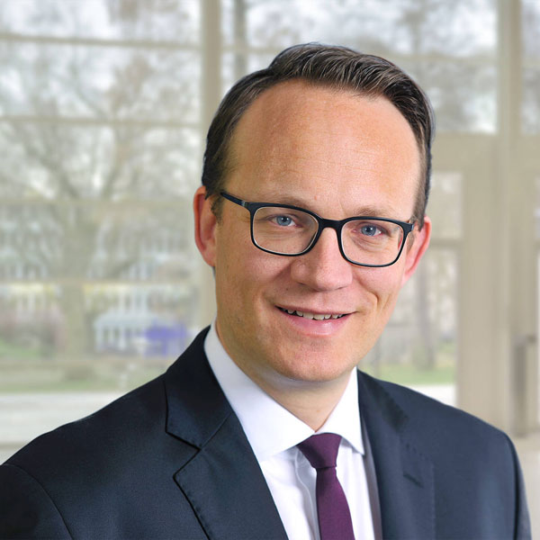 Ein Mann trägt einen Anzug mit einer Krawatte und steht in einem modernen, hellen Raum mit Fenstern im Hintergrund, Portrait von Dr. Markus Krebber.