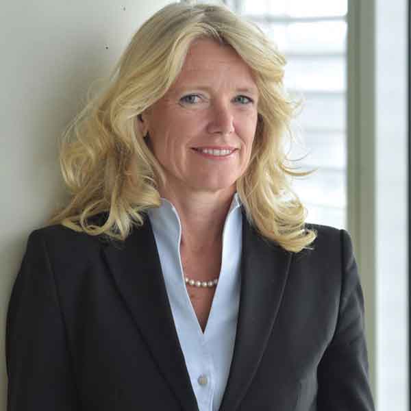 Eine Frau mit blonden Locken trägt einen schwarzen Blazer über einem hellblauen Hemd. Sie steht neben einem Fenster, Portrait von Stephanie Schunck.