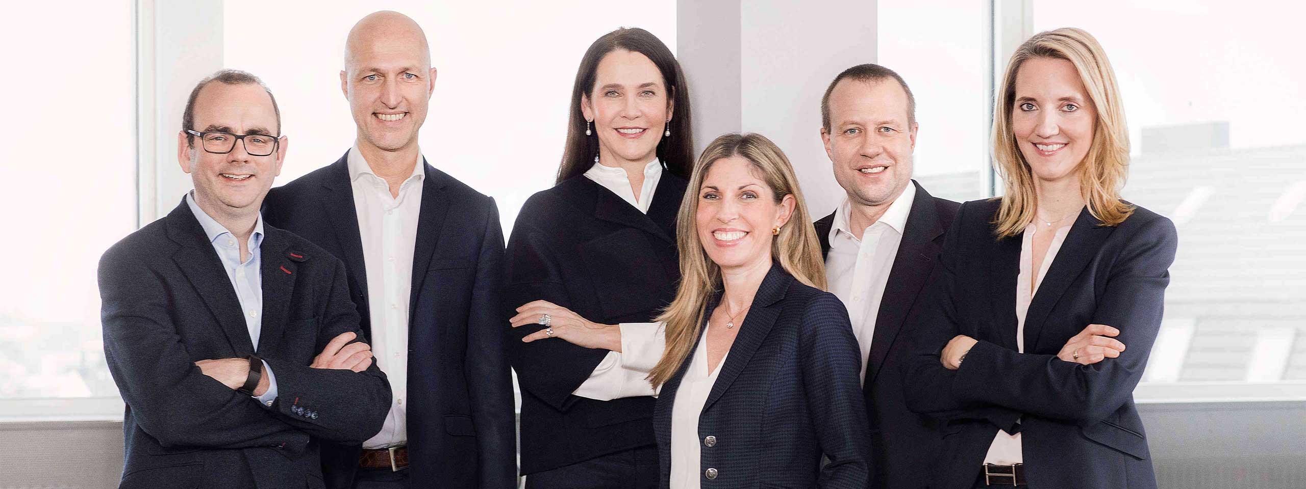 A group of six individuals in professional attire, standing against large windows with natural light.