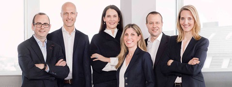 A group of six individuals in professional attire, standing against large windows with natural light.