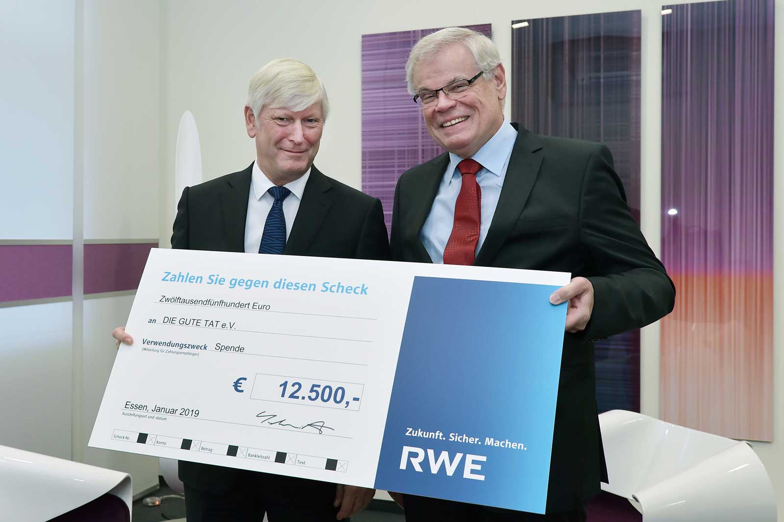 Zwei Personen halten einen großen Scheck über 12.500 Euro in einem modernen Büro.