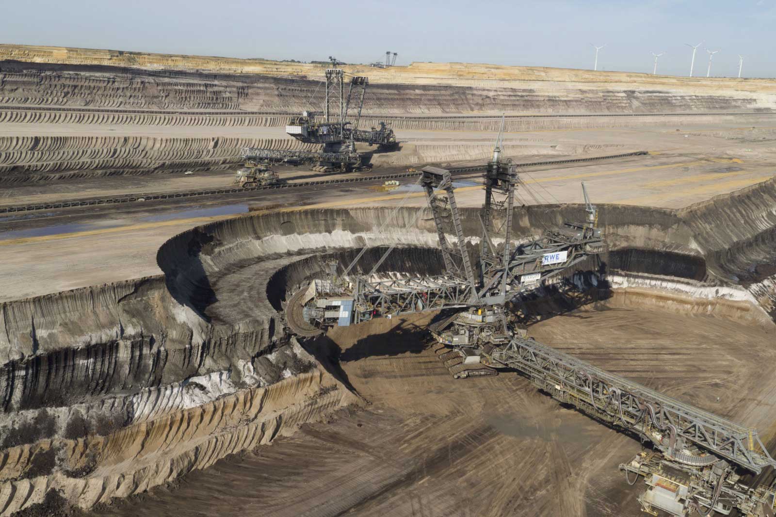 Ein Tagebau mit großen Maschinen, die Braunkohle abbauen. Der Himmel ist klar und Windkraftanlagen sind im Hintergrund sichtbar.