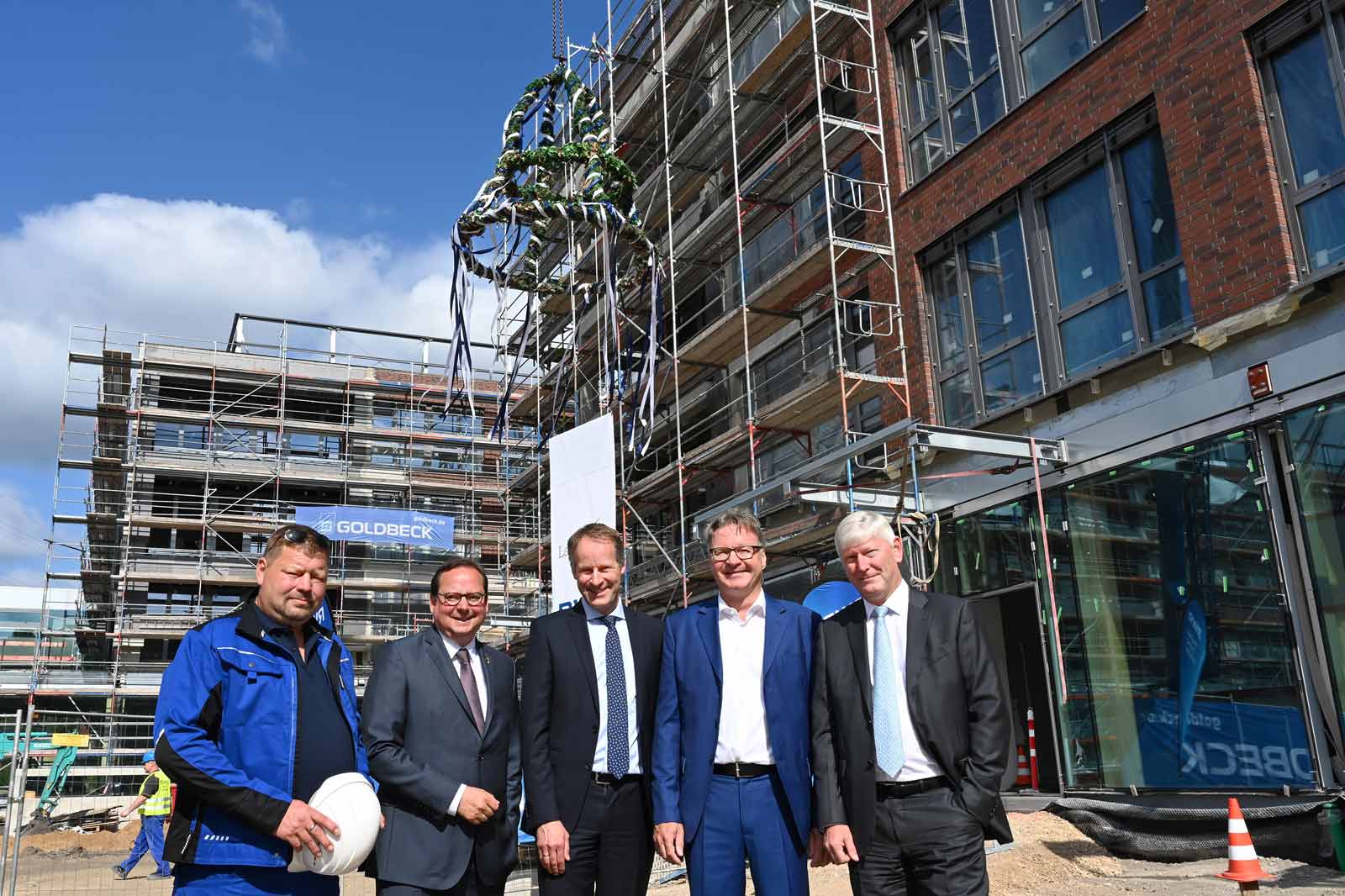 Five individuals stand in front of a construction site with scaffolding and a Goldbeck logo visible.