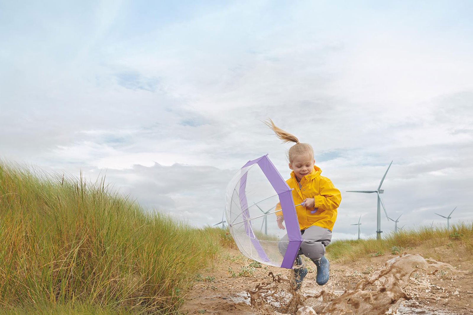 A child in a yellow raincoat joyfully jumps with a purple umbrella through puddles on a grassy field.