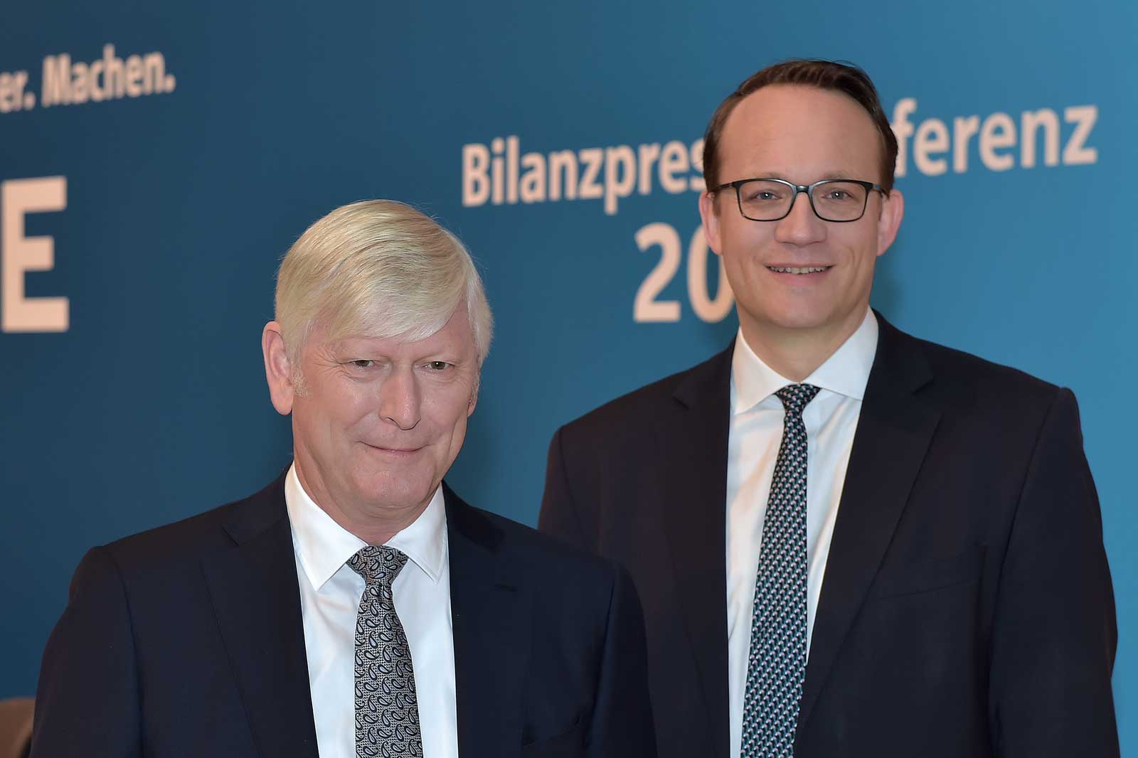 Two men in formal attire stand against a blue background featuring text about a balance sheet press conference.