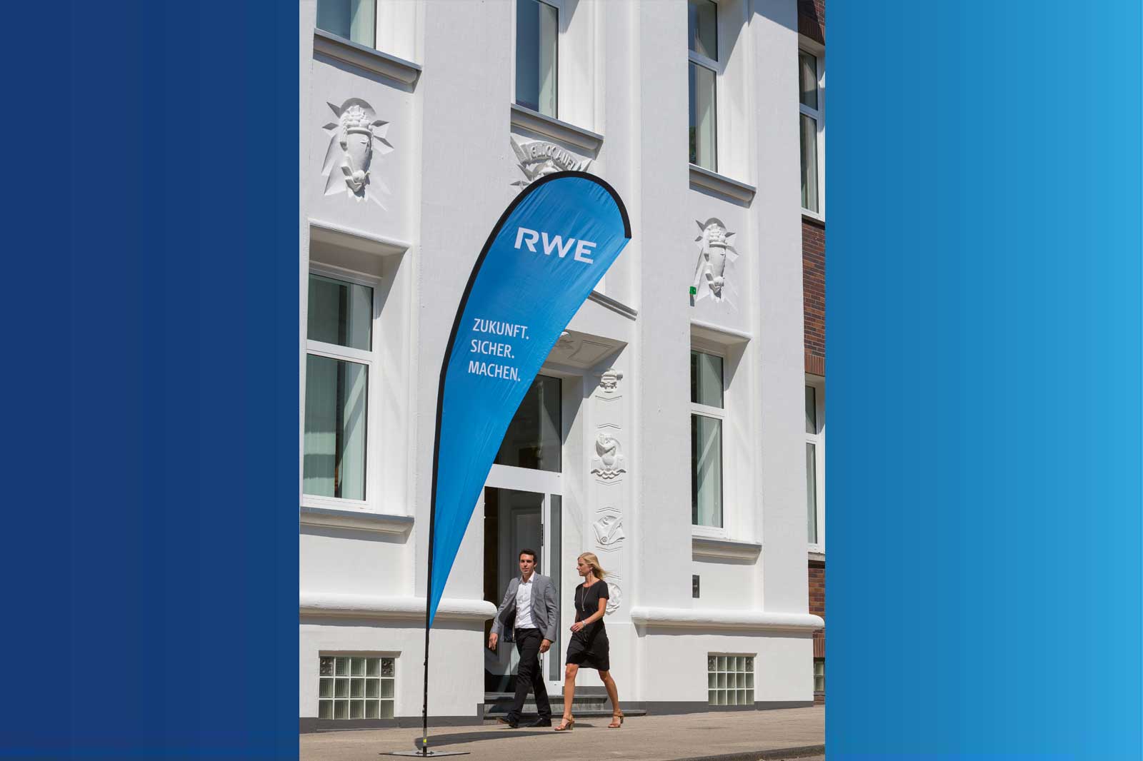 Two people exiting a building with a blue flag displaying RWE, sunlight illuminates the white facade.