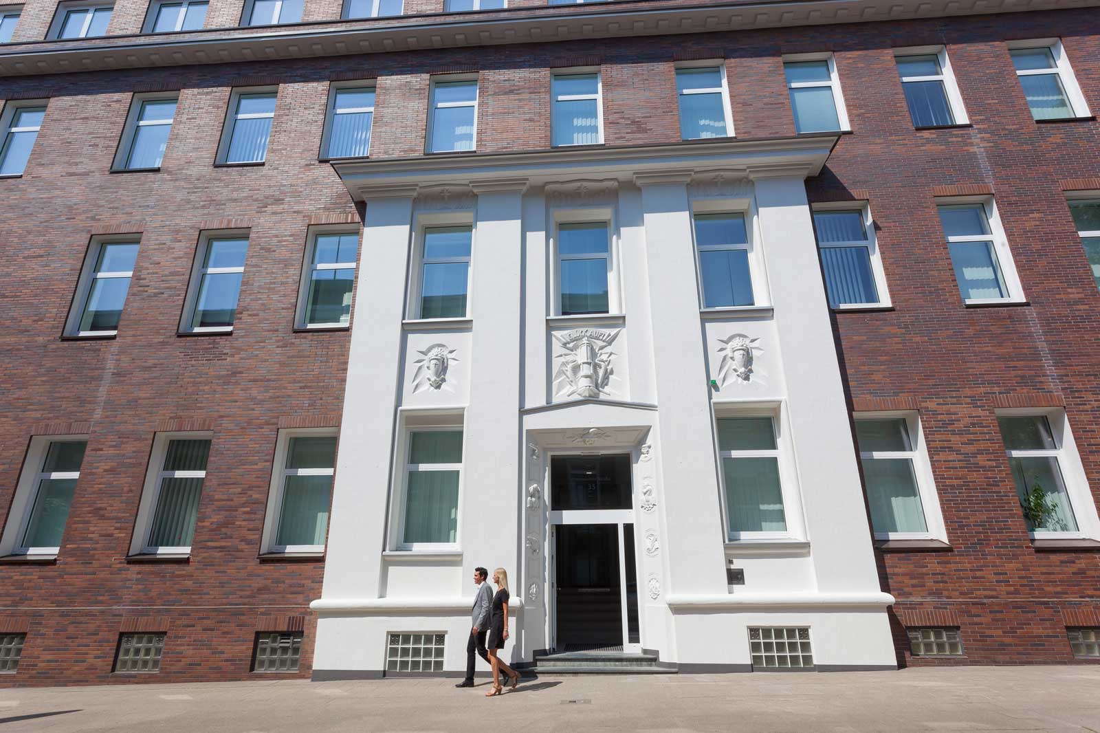 Two people walk past a modern, multi-storey building with red bricks and white decorative elements.