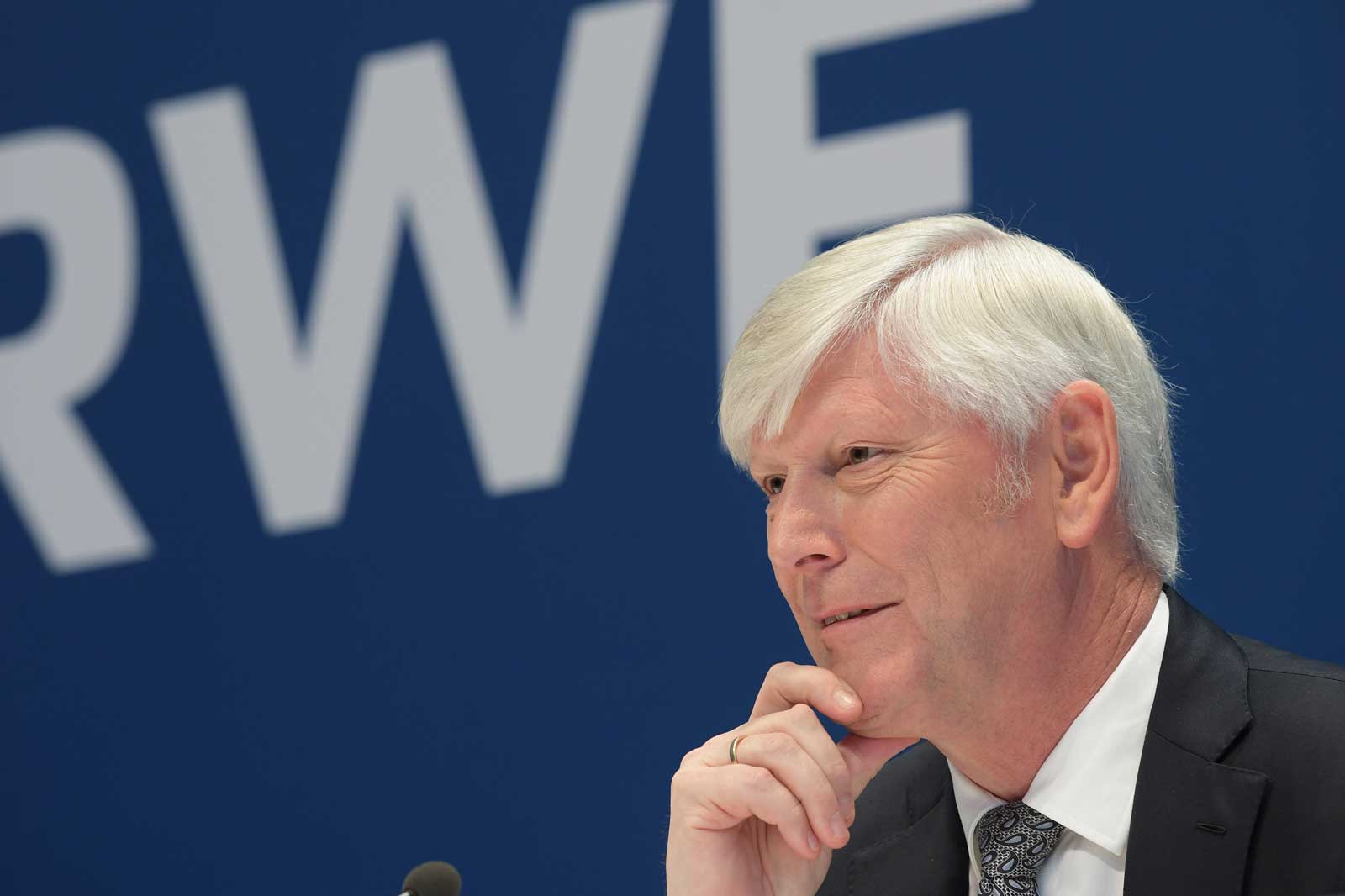 A man in a suit sits at a table, thoughtfully gazing into the camera, with a large RWE sign in the background.
