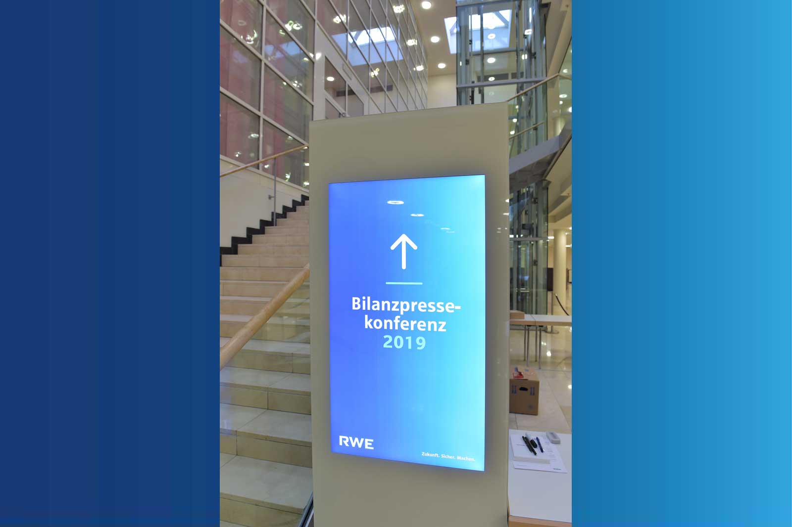 A digital sign displays 'Bilanzpressekonferenz 2019' in a modern building with a staircase in the background.