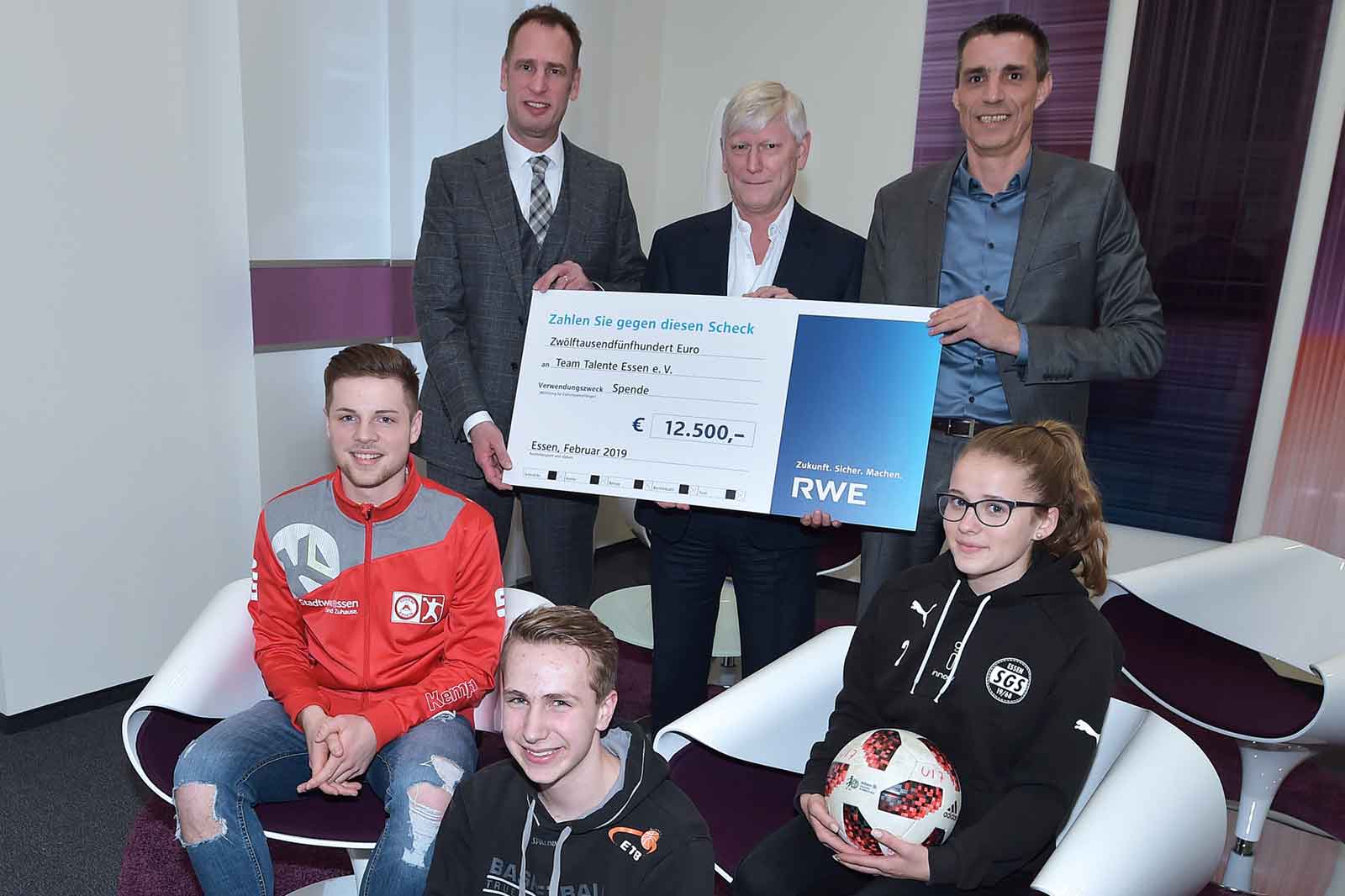 Eine Gruppe von Menschen hält einen großen Scheck über 12.500 Euro für ein Team in Essen. Es gibt einen Fussball auf dem Boden.