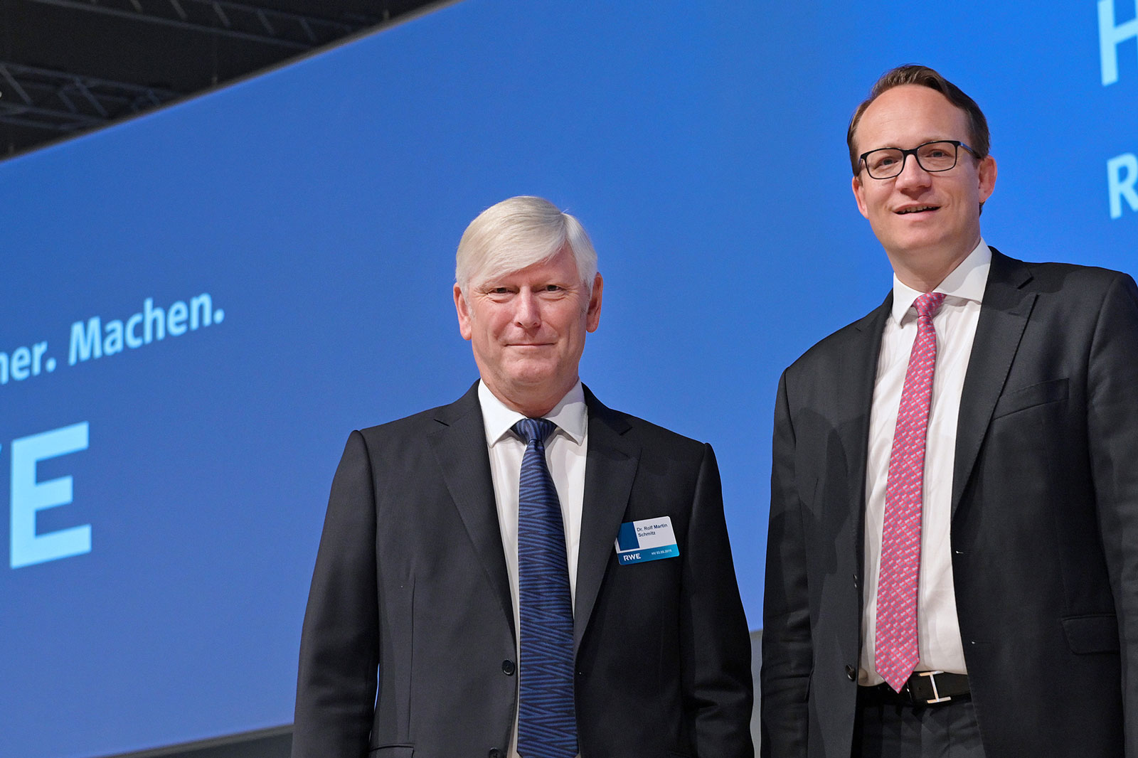 Two men in suits stand together in front of a blue background with text. The left one wears a tie and a name badge.