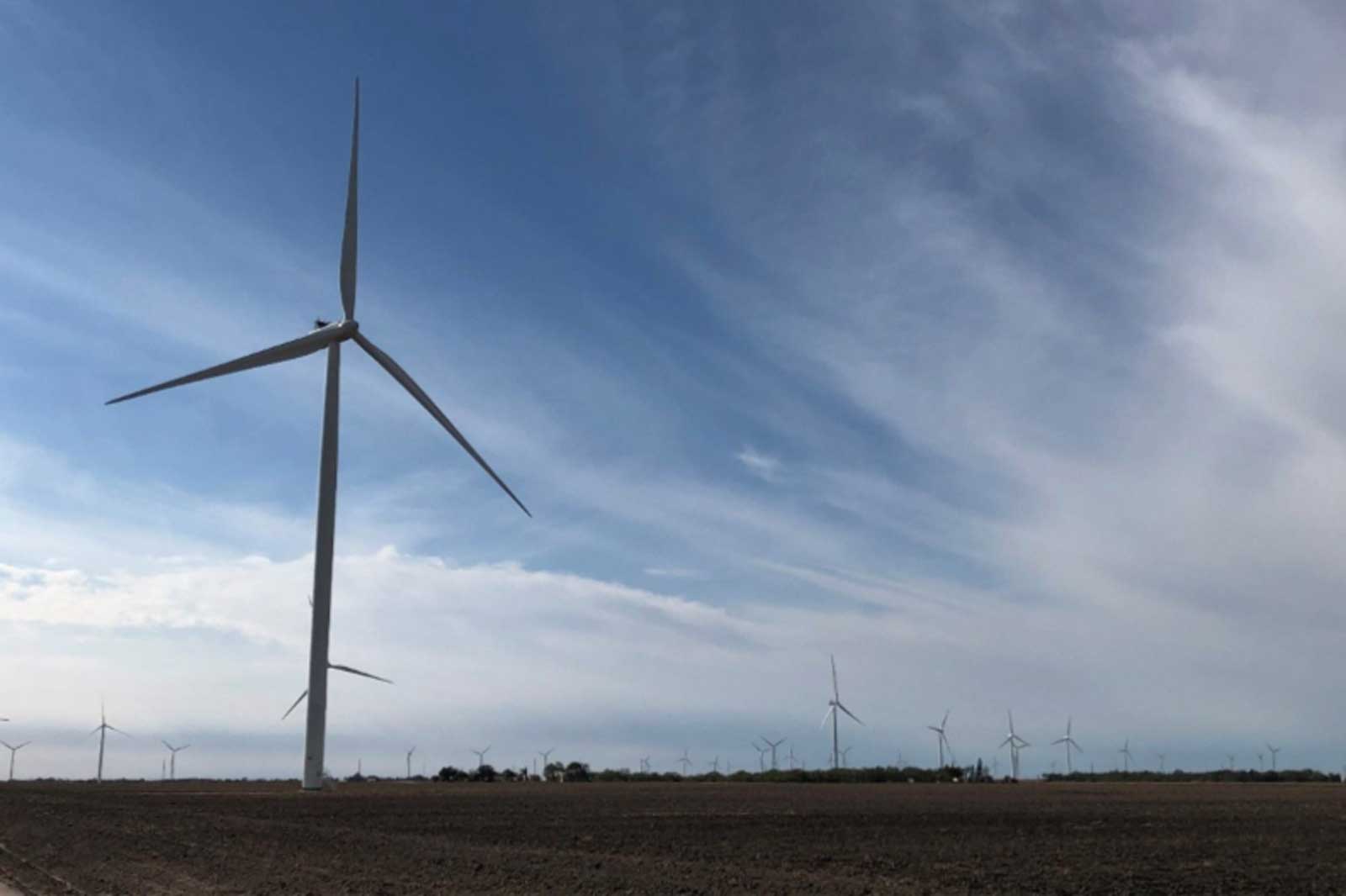 A wind farm with numerous wind turbines under a blue sky and wispy clouds over brown, tilled land.