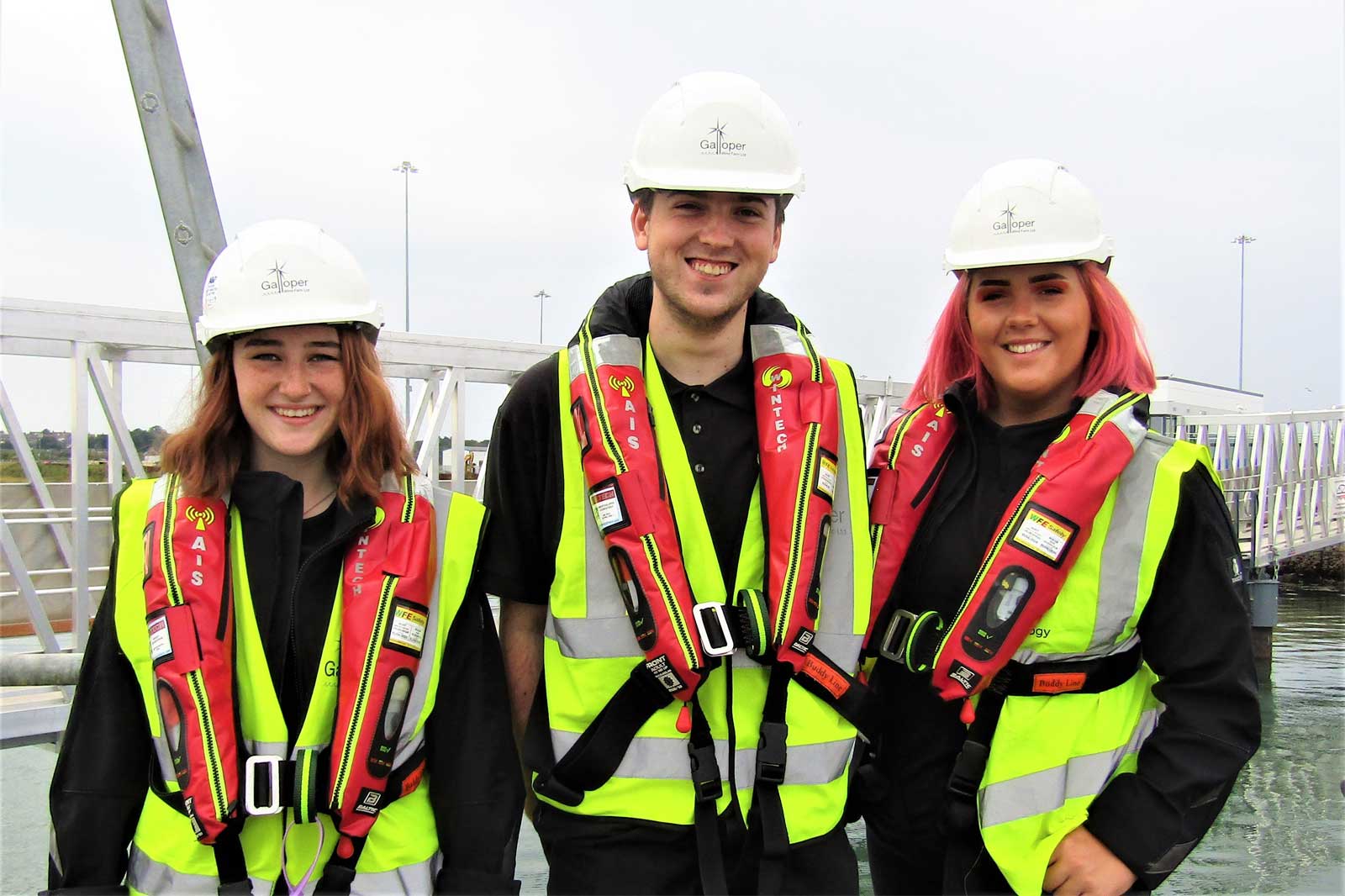 Three individuals stand on a dock wearing bright yellow and black safety vests and white helmets. They are ready for a maritime activity.
