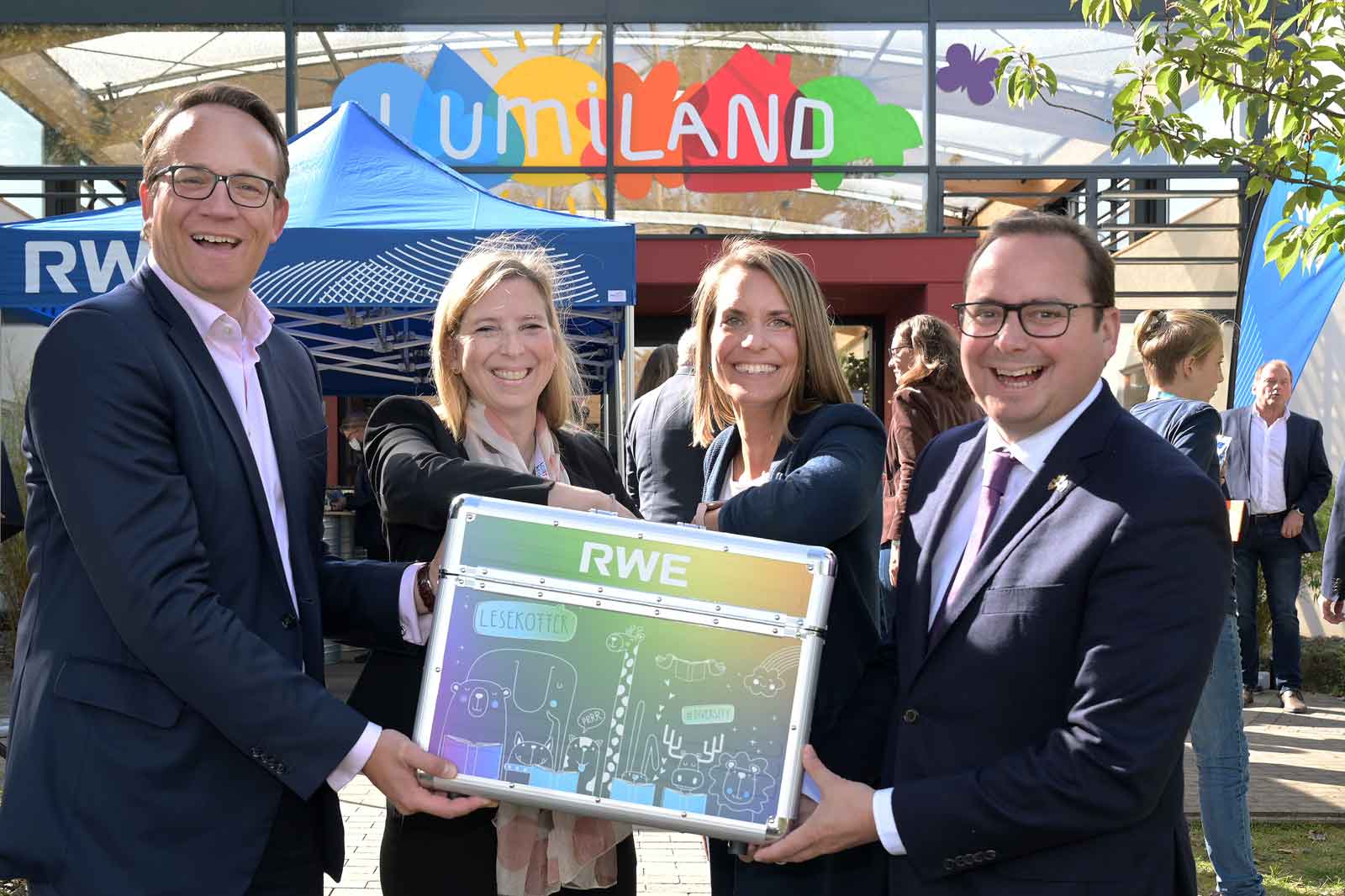 Gruppenfoto vor dem Lumiland, Personen halten einen bunten Koffer mit RWE-Logo, im Hintergrund Zelte und Menschen.
