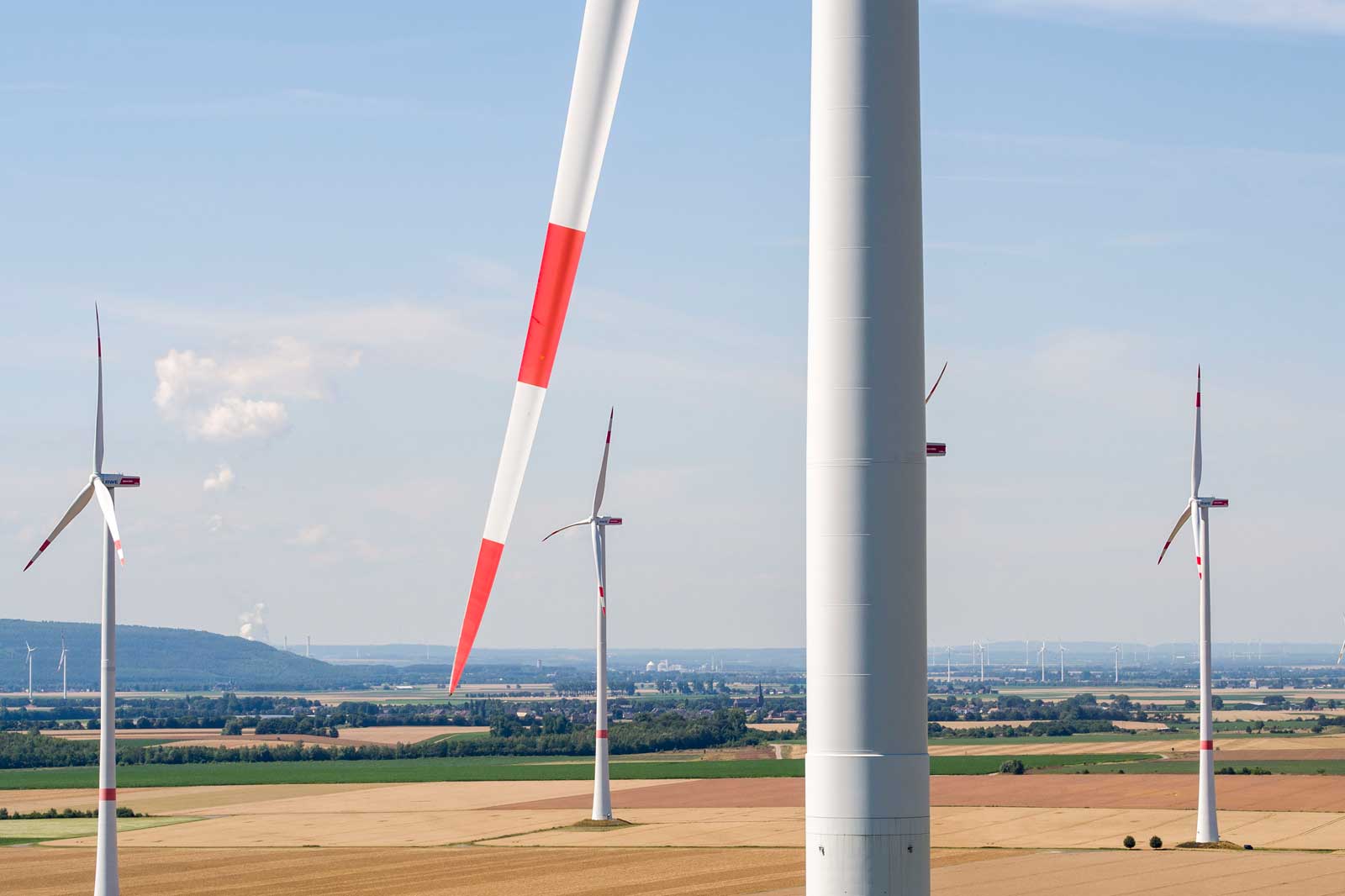 A panoramic view of multiple wind turbines with red and white blades against a clear blue sky and fields in the background.