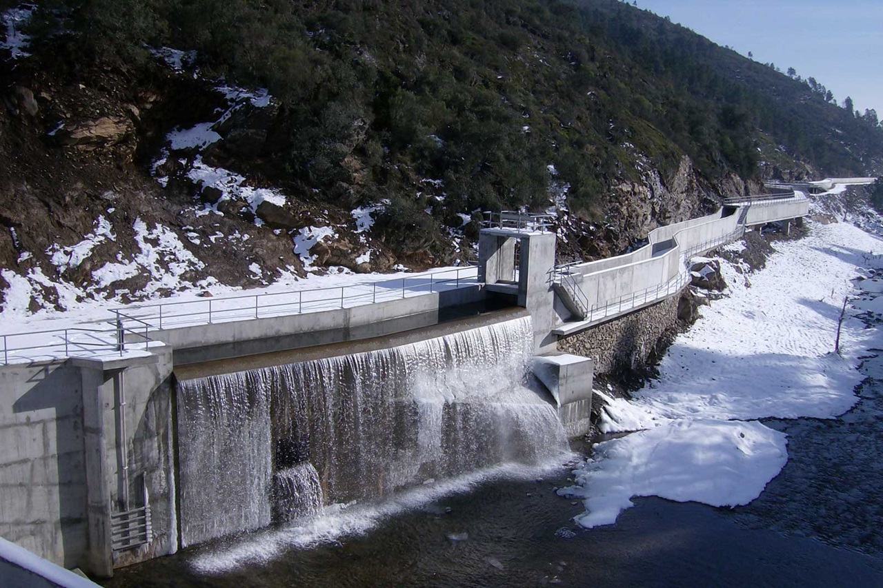 A dam with flowing water and snow on the bank, surrounded by rocks and trees.