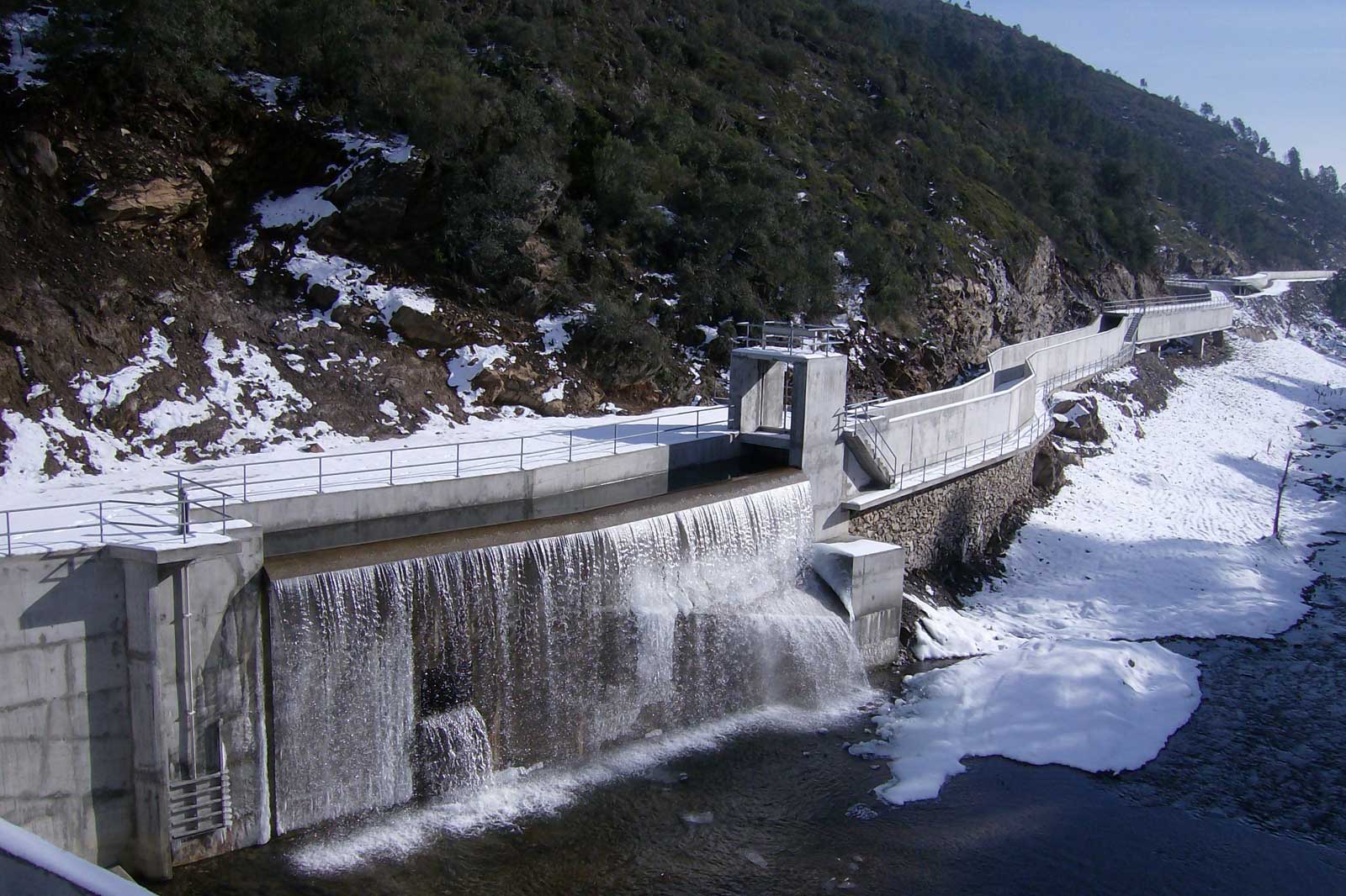 Ein Staudamm mit fließendem Wasser und Schnee am Ufer, umgeben von Felsen und Bäumen.