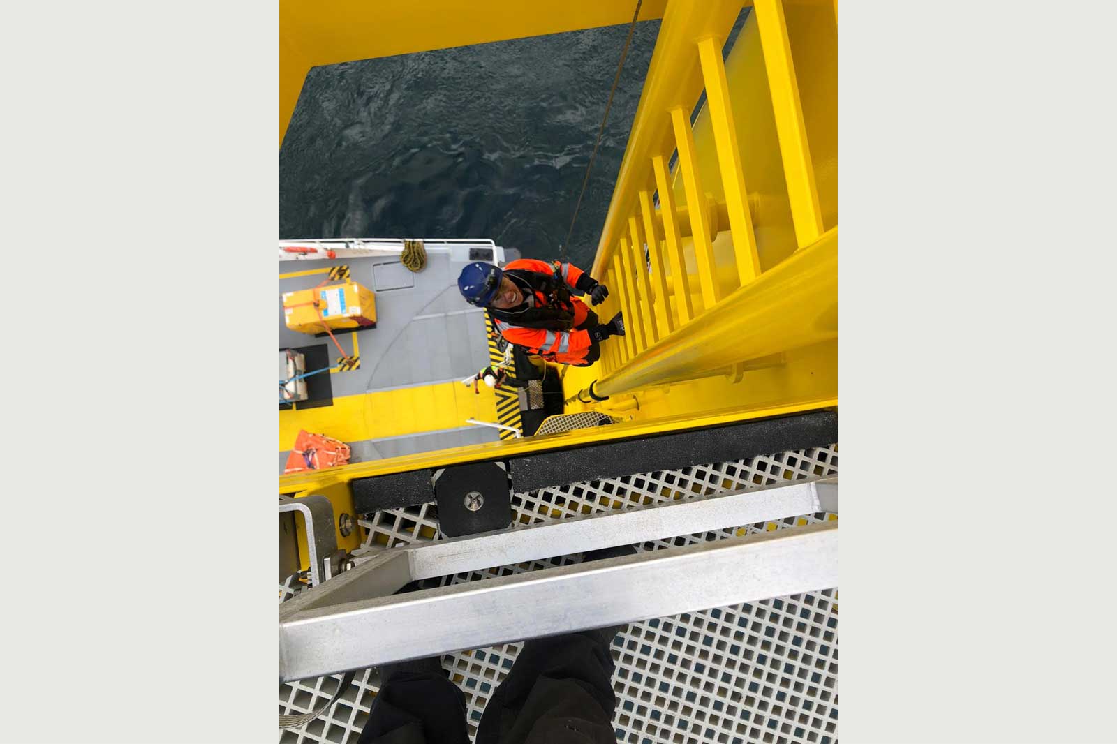 A worker in safety gear climbs a yellow ladder from a vessel, with a view of the sea and deck below.