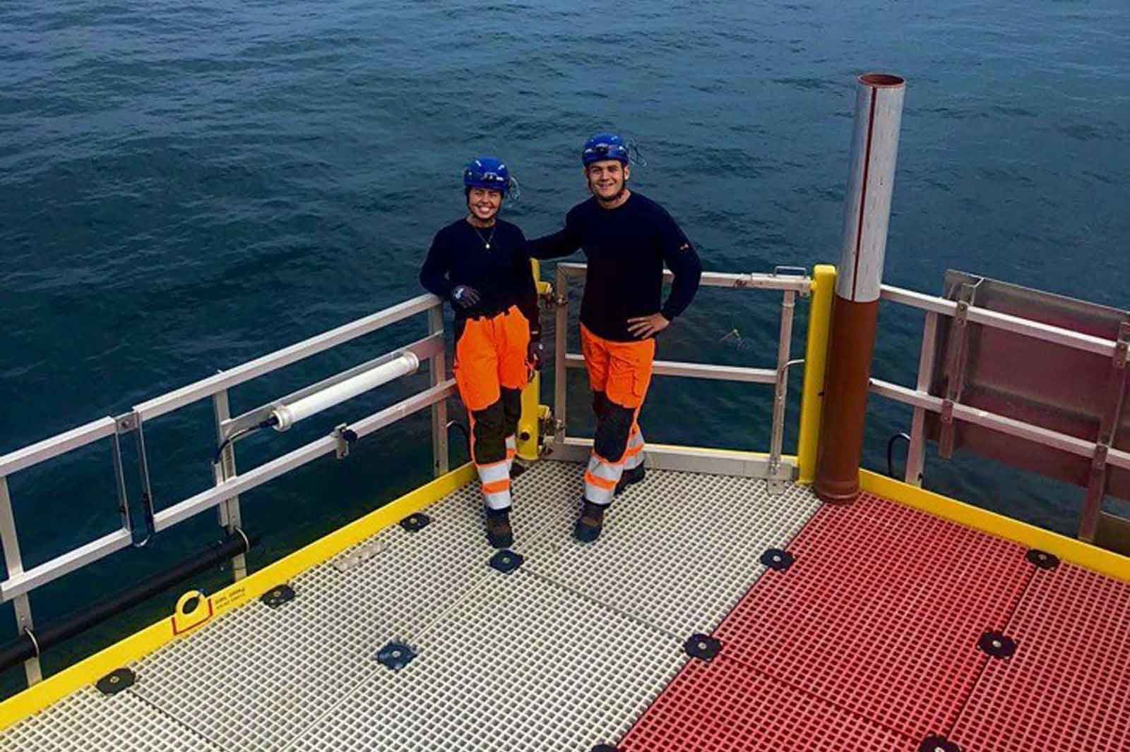 Two workers in safety gear pose on a platform at sea, surrounded by blue water and a safety railing.