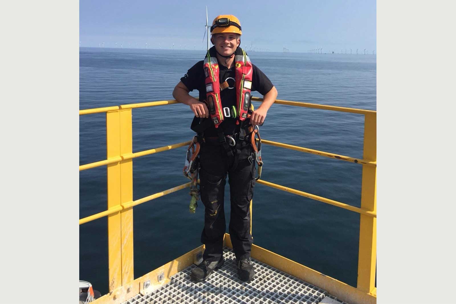 A safety worker stands on a yellow platform by the sea, wearing a harness and life jacket, with wind turbines in the background.