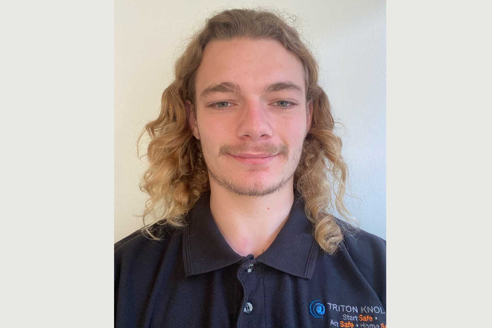 A person wearing a black polo shirt with a logo, featuring curly hair, against a light background.
