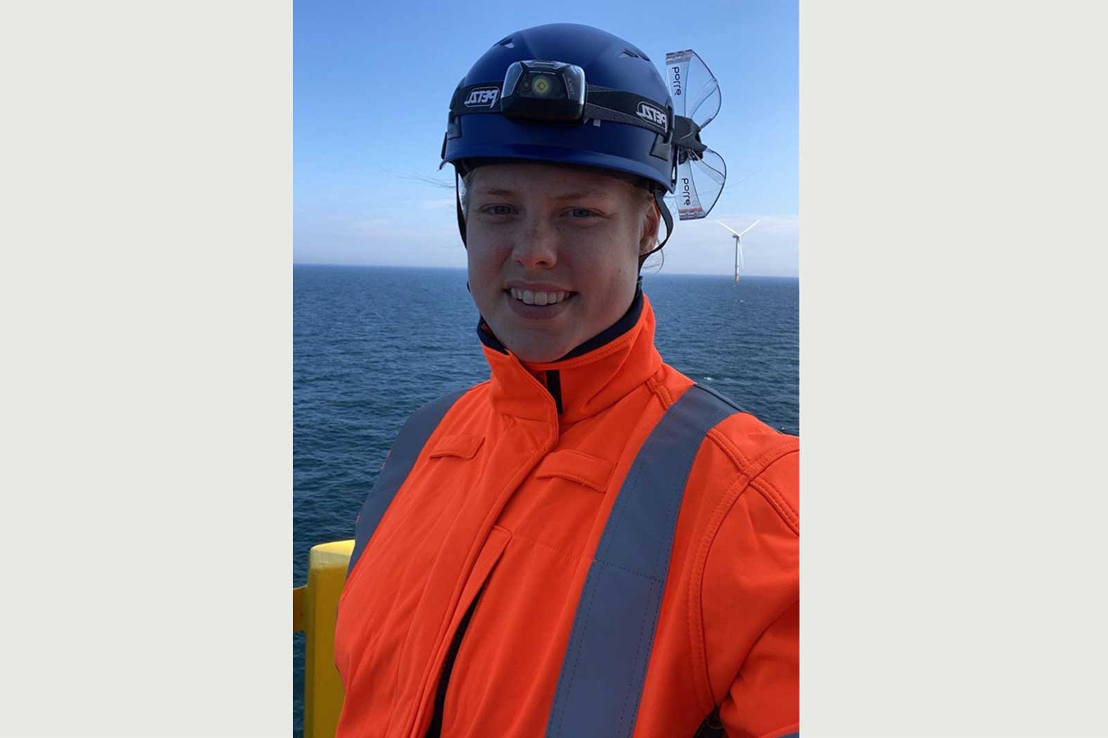 A person in an orange safety jacket and helmet poses at sea with wind turbines visible in the background.
