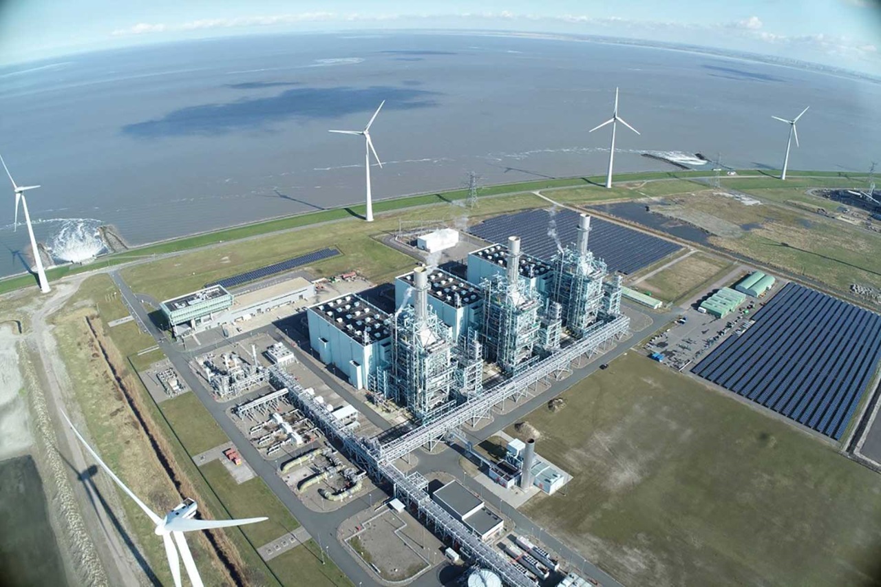 Aerial view of an energy park featuring a gas power plant, wind turbines, and solar panels beside the water.