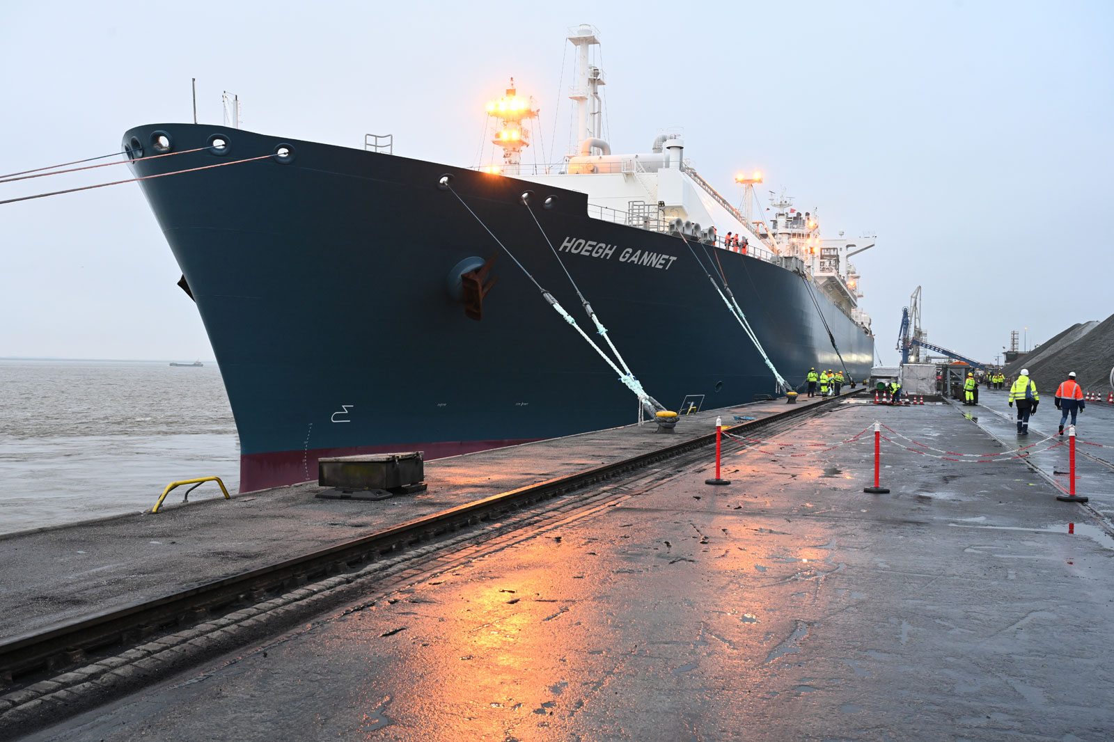 A large ship is docked at a quay. Workers in safety gear are active, with a crane and cargo in the background.