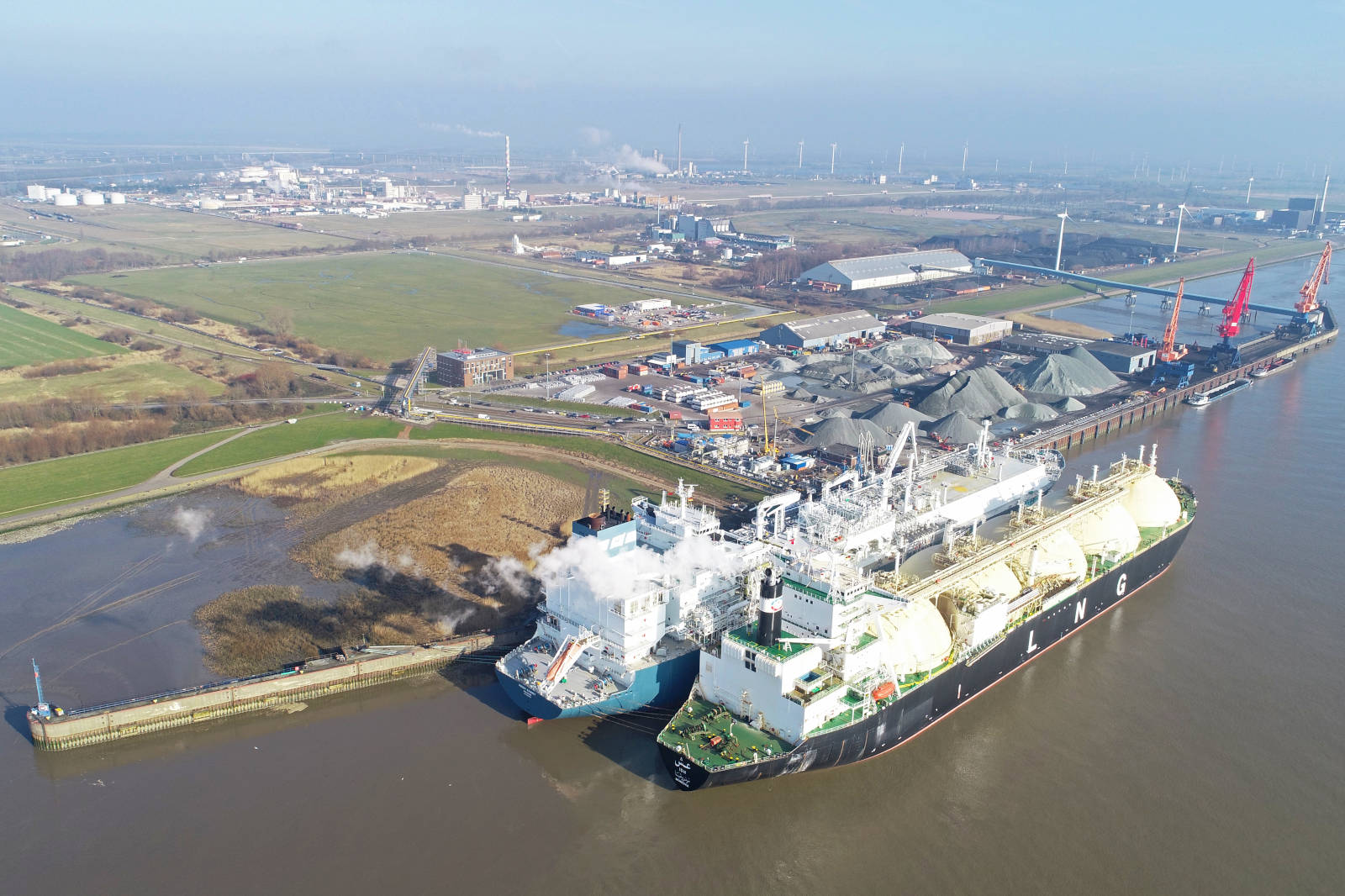 Luftaufnahme eines LNG-Schiffs im Hafen, umgeben von Industrie und grünen Flächen, mit rauchenden Schornsteinen im Hintergrund.