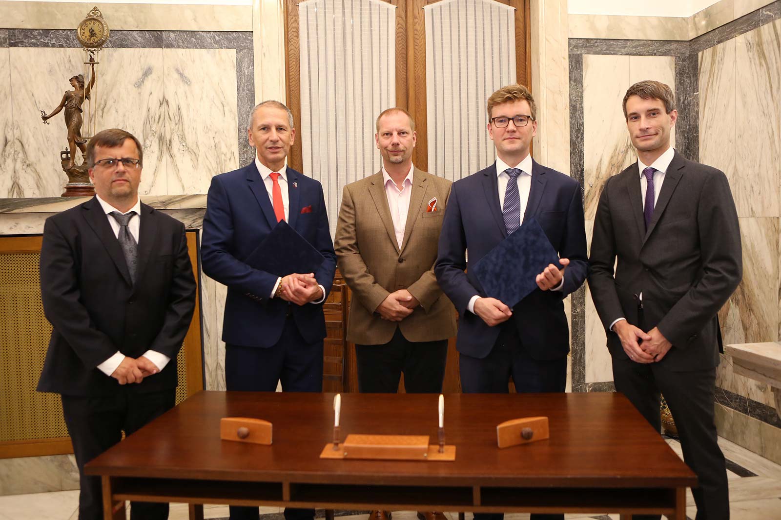Fünf Männer in Anzügen stehen vor einem Tisch in einem eleganten Raum mit einer Statue und Marmormauern.