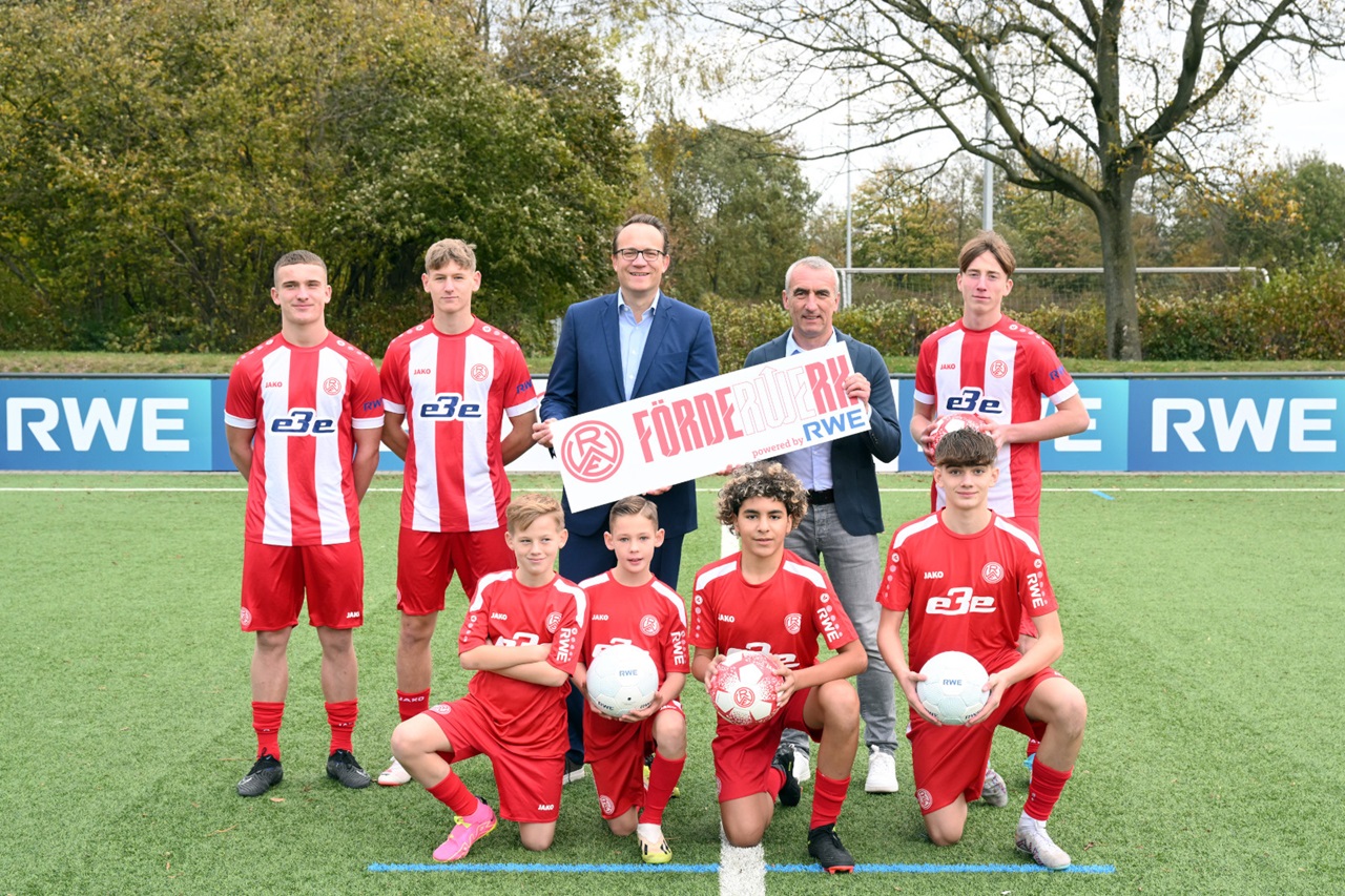Eine Gruppe junger Fußballspieler in roten Trikots posiert mit einem Schild auf dem Platz und zeigt Teamgeist.
