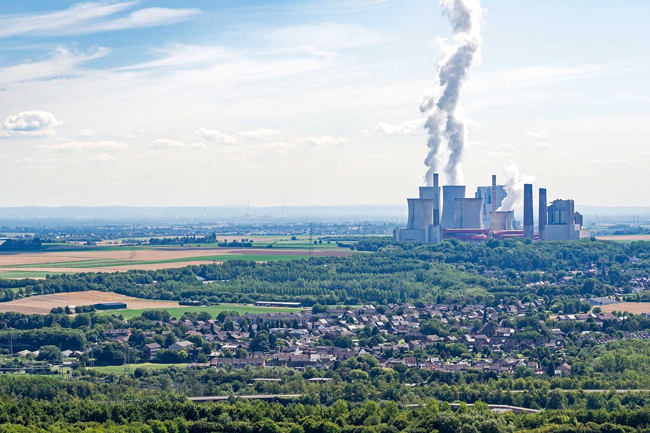 Eine weitläufige Landschaft mit einem Kraftwerk im Hintergrund und rauchenden Kühltürmen, umgeben von Feldern und Bäumen.
