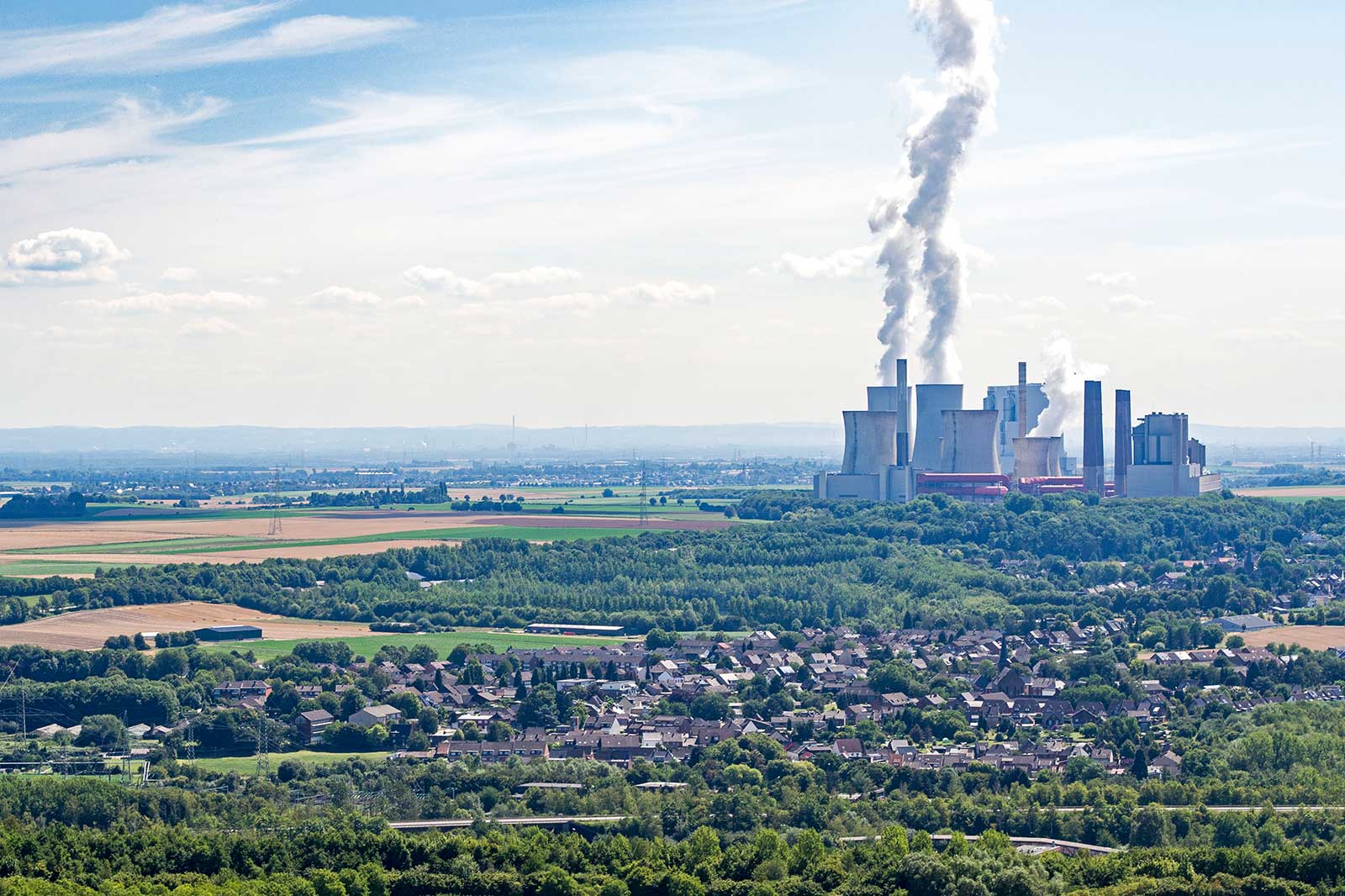 Eine weitläufige Landschaft mit einem Kraftwerk im Hintergrund und rauchenden Kühltürmen, umgeben von Feldern und Bäumen.