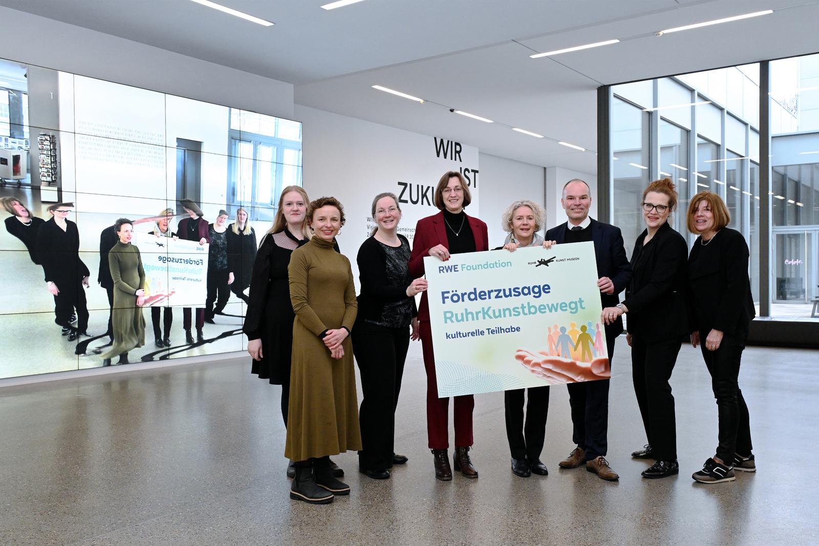 Gruppe von Personen hält ein Schild mit der Aufschrift 'Förderzusage RuhrKunstbewegt' in einer modernen Umgebung.