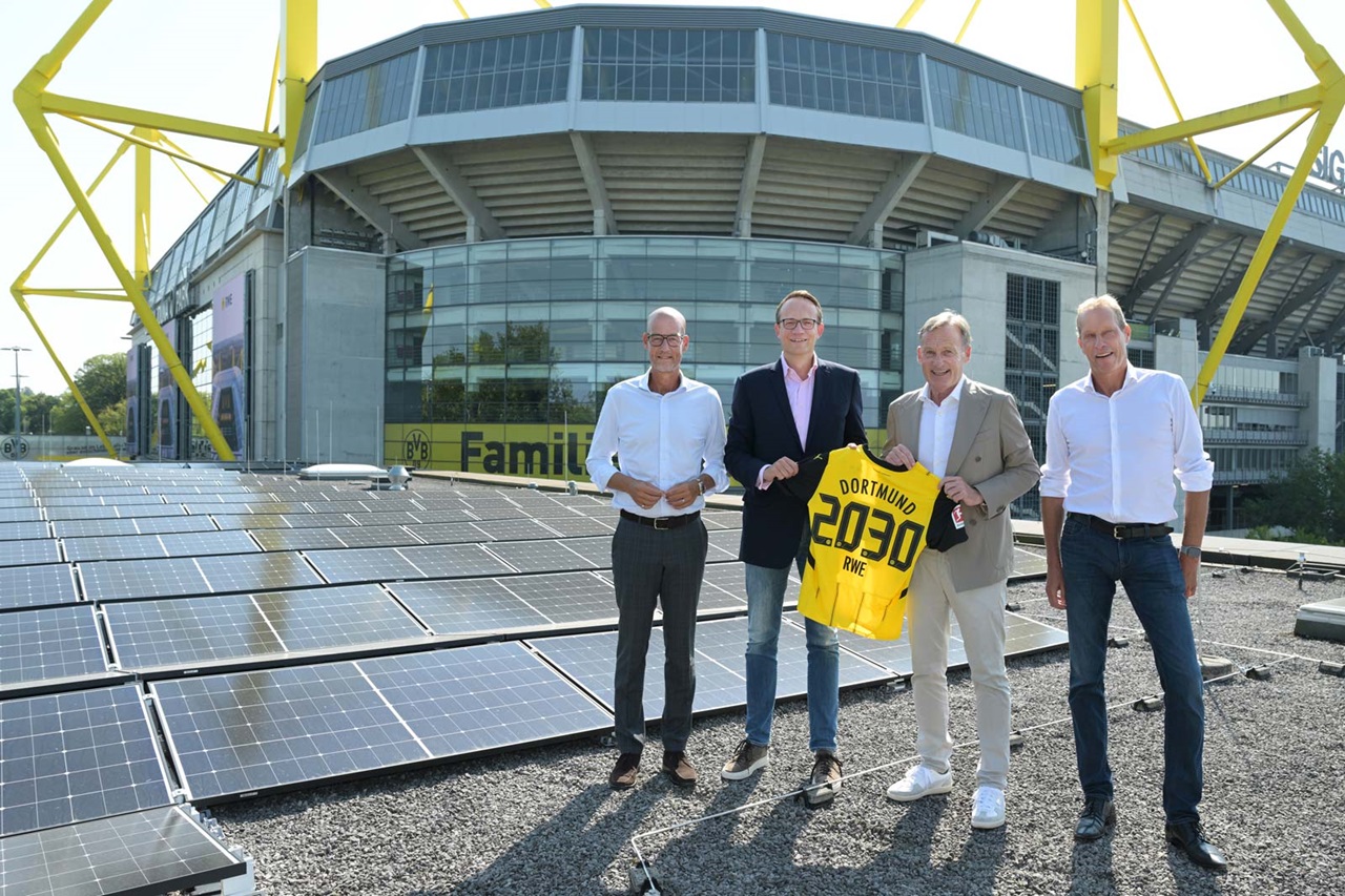 Vier Personen stehen auf einem mit Solarpaneelen bedeckten Dach und halten ein gelbes Trikot mit 'Dortmund 2030 RWE', während ein Stadion im Hintergrund zu sehen ist.