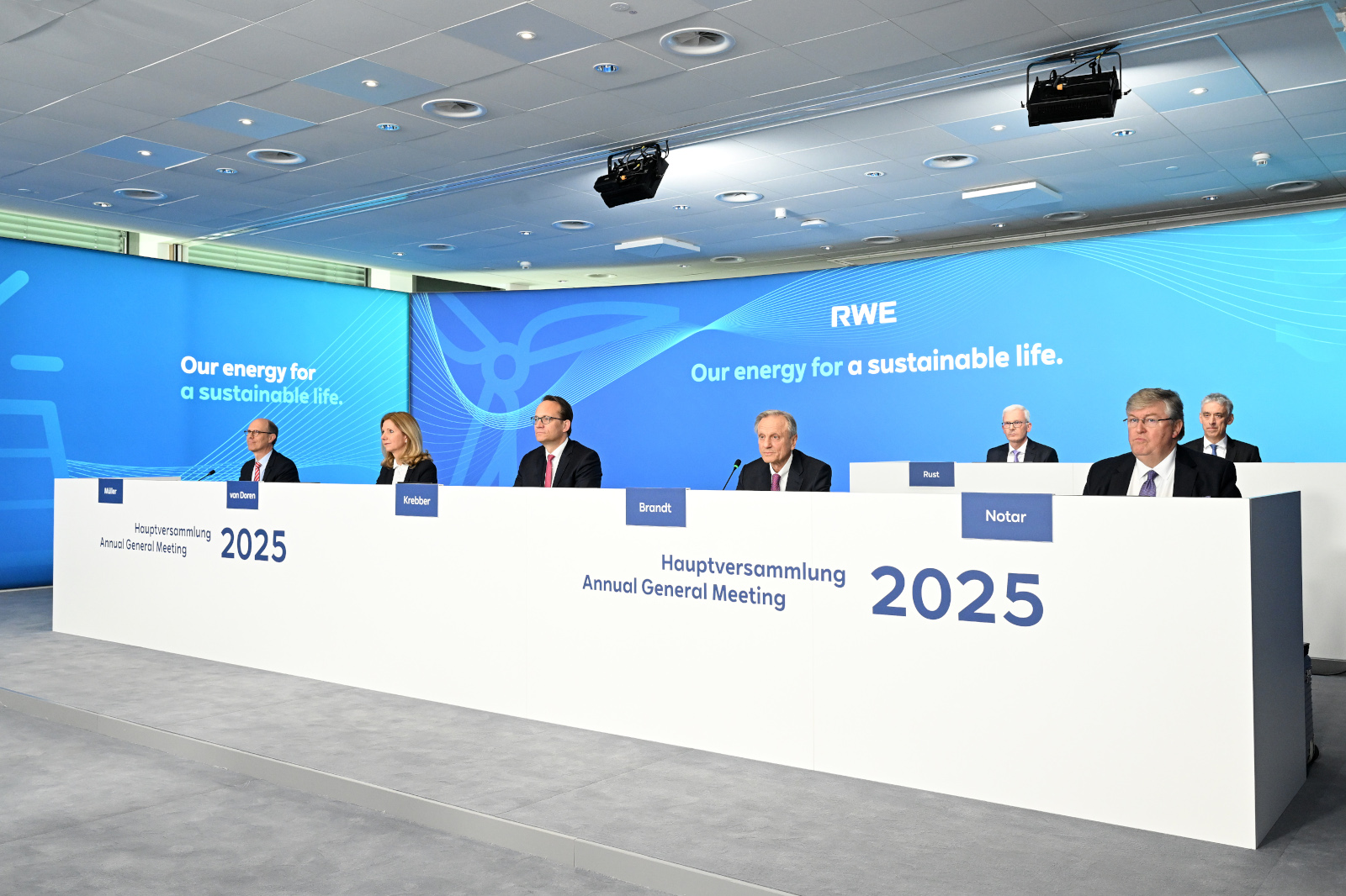 Die Vorstandmitglieder von RWE sitzen an einem Tisch für die Hauptversammlung 2025, mit Hintergrundgrafik zur nachhaltigen Energie.